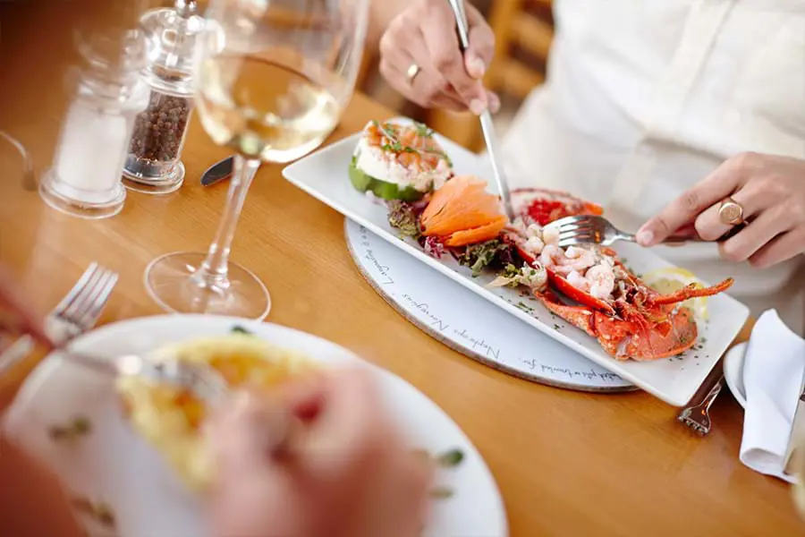 Image of the table with two people having dinner, from the point of view from behind one's shoulder, the plate in the forefront is blurry but the other shows a plate of lobster and vegetables, and the persons' two hands cutting it up with a knife and fork. A glass of white wine and salt and pepper grinders are also on the table.