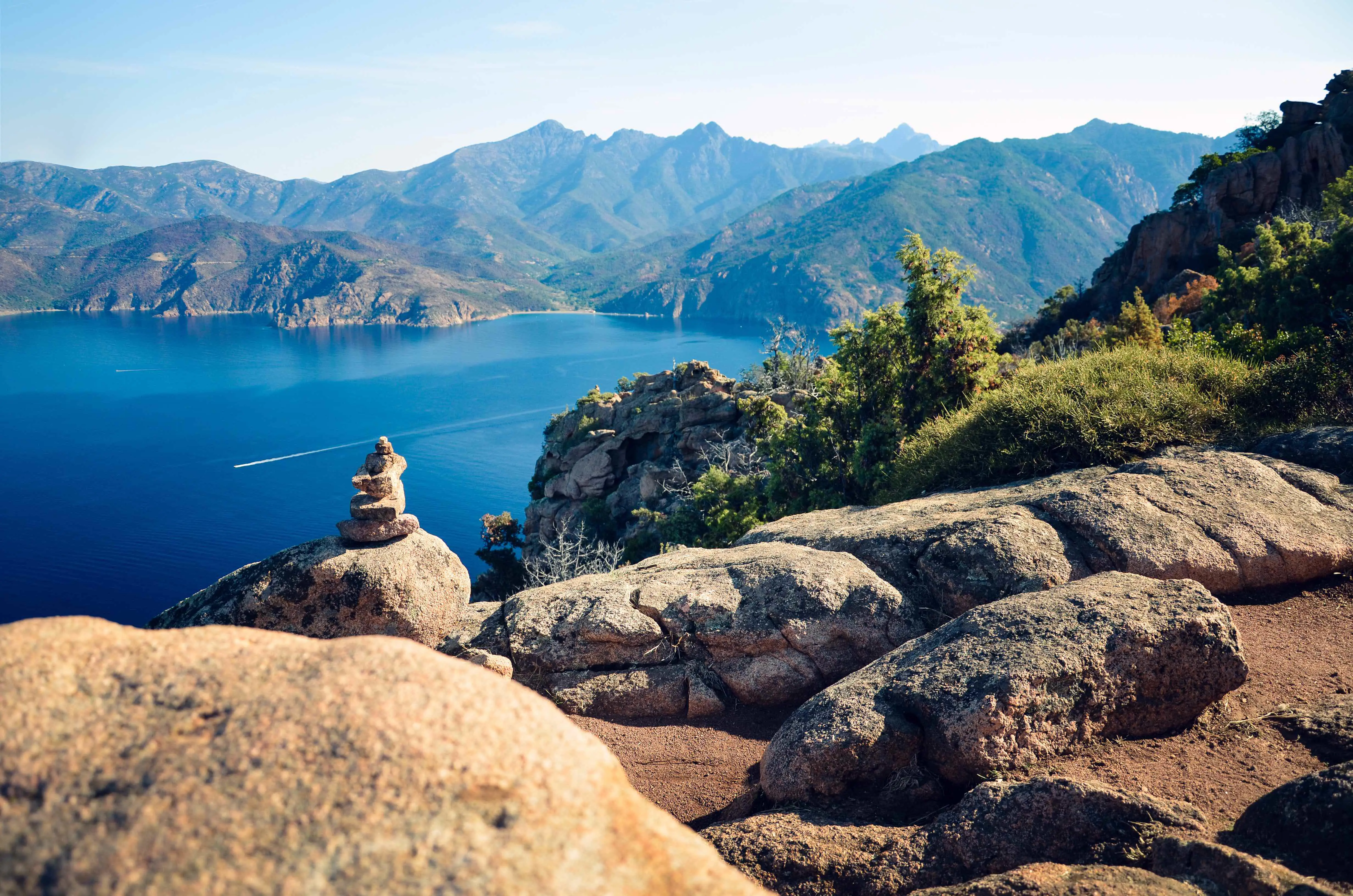 View of the sea with rocks in the forefront and a bush to the right. Mountains in the distance on the other side of the water