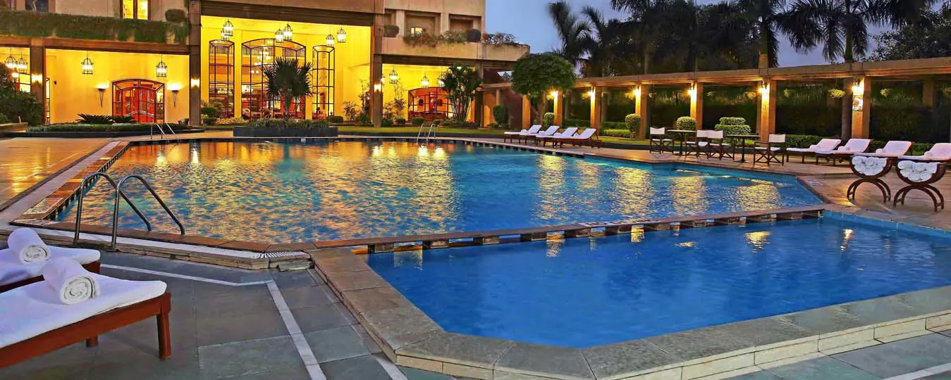 Illuminated swimming pool surrounded by sun loungers and palm trees at Eros Hotel, New Delhi, with the hotel’s lit-up façade in the background