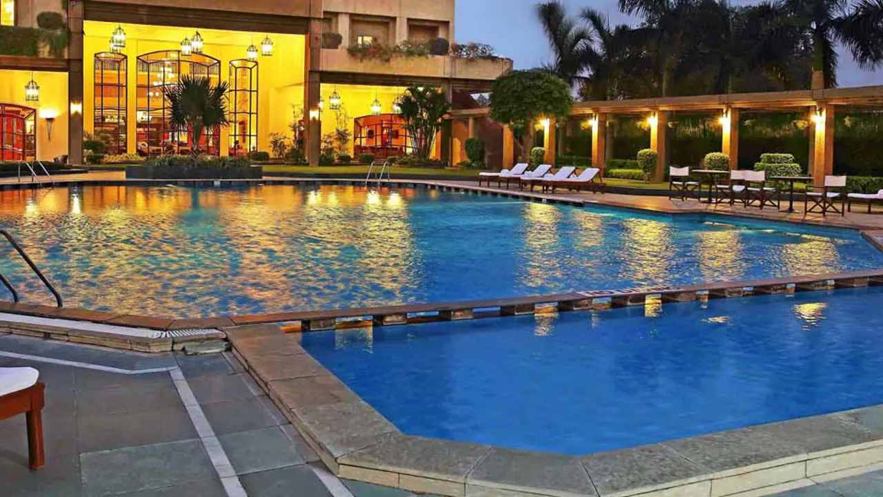 Illuminated swimming pool surrounded by sun loungers and palm trees at Eros Hotel, New Delhi, with the hotel’s lit-up façade in the background