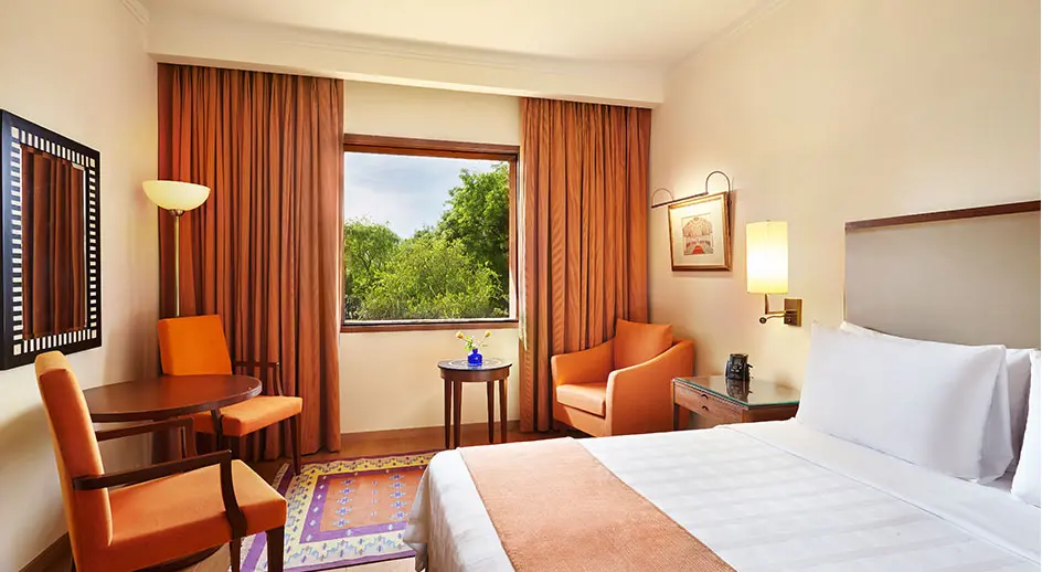 A double room at the Trident hotel in Agra featuring warmly lit décor and furnishings and white linens on a double bed. The room has a view of the surrounding landscape.