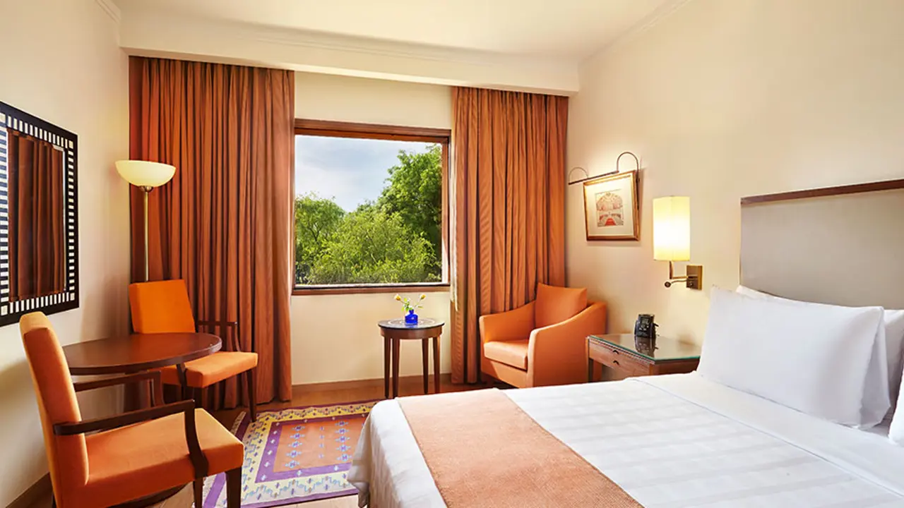 A double room at the Trident hotel in Agra featuring warmly lit décor and furnishings and white linens on a double bed. The room has a view of the surrounding landscape.