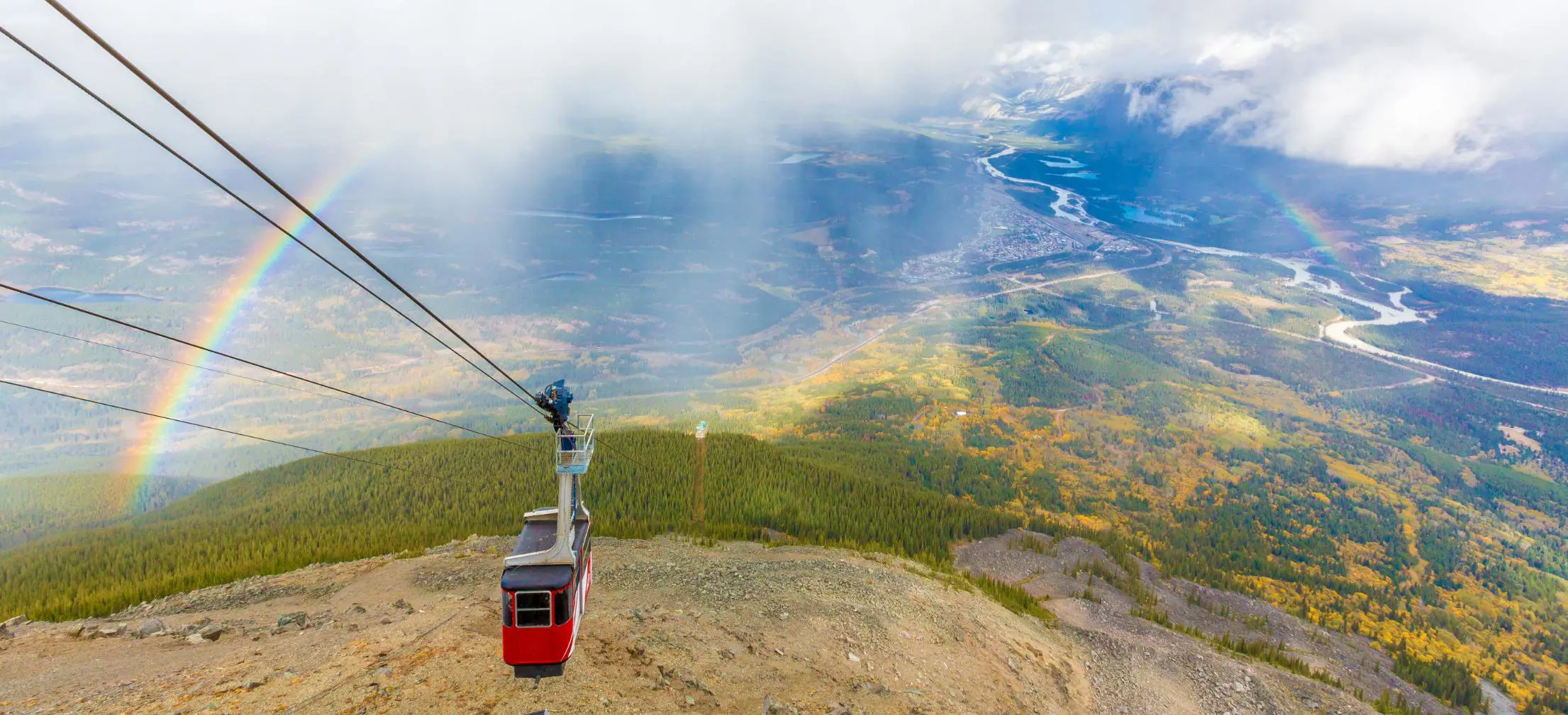 View of a cable car over Jasper National Park in Alberta, Canada, showing forested slopes and rugged mountain peaks beneath a rainbow arching across the sky