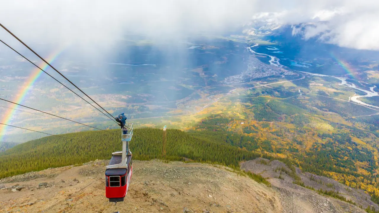 View of a cable car over Jasper National Park in Alberta, Canada, showing forested slopes and rugged mountain peaks beneath a rainbow arching across the sky
