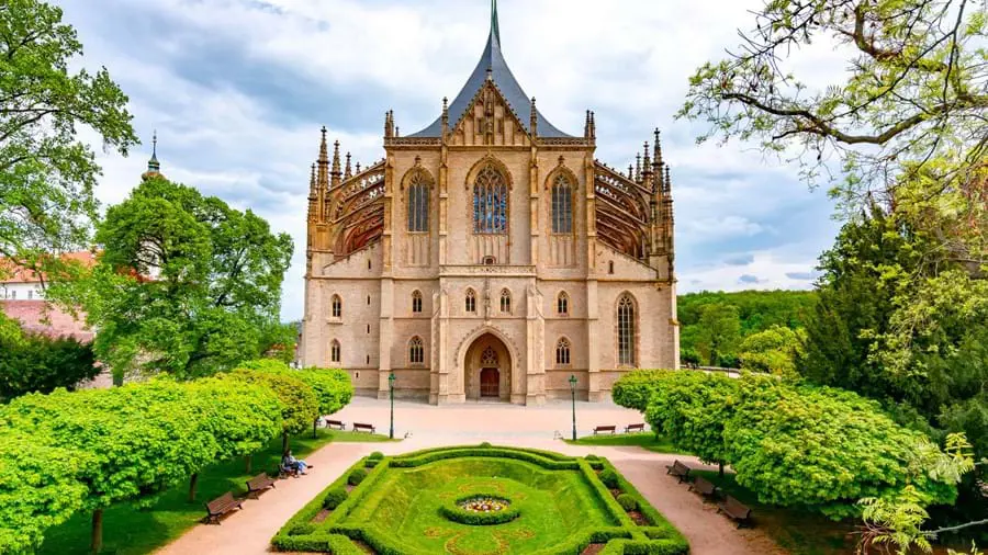 A beige coloured church with a pointed roof and small turrets on either side. A lawn in front of it and bushes either side.