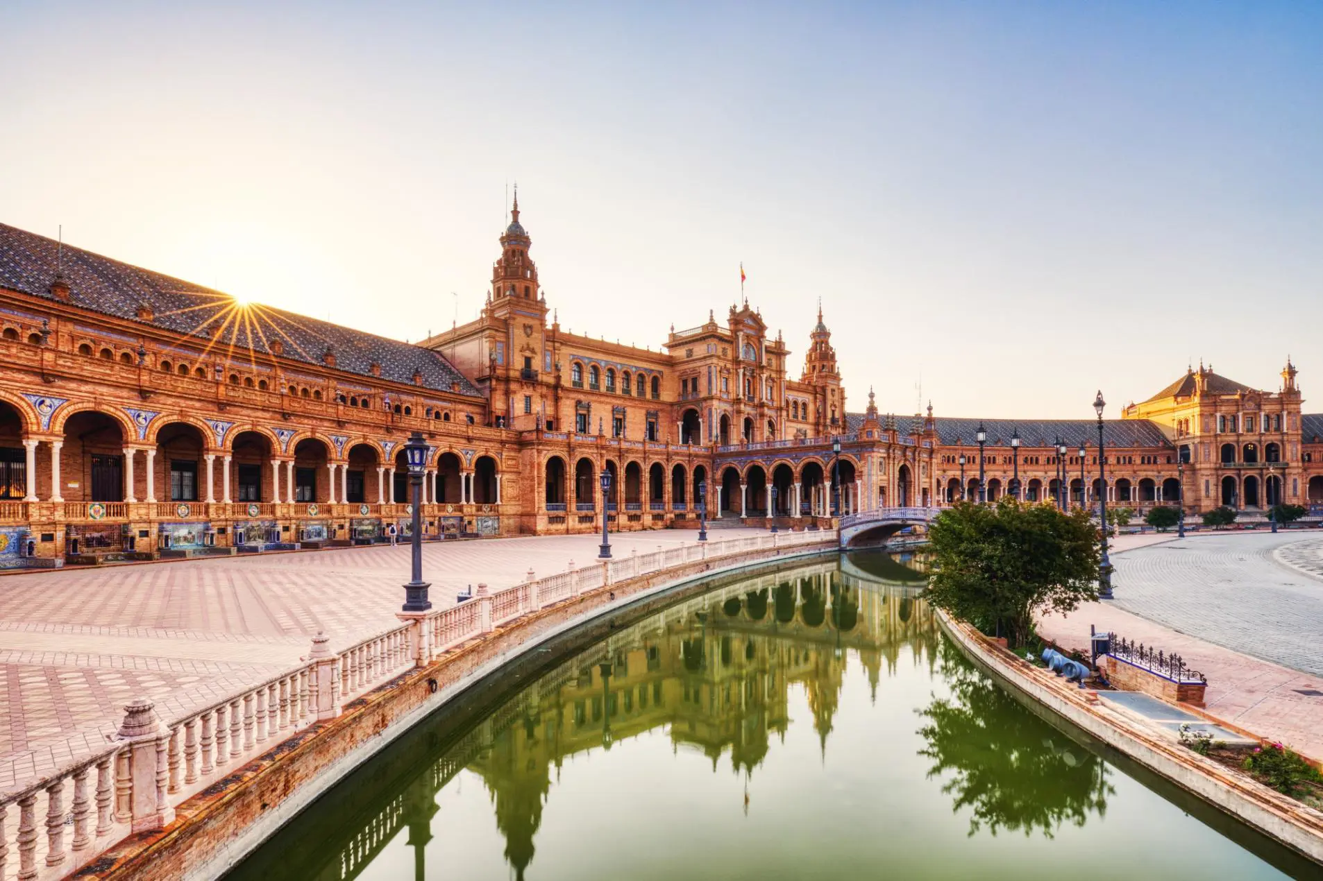 Wide view of Plaza de España in Seville, featuring its grand semicircular building, ornate tilework, and the small river with a bridge crossing it in the distance