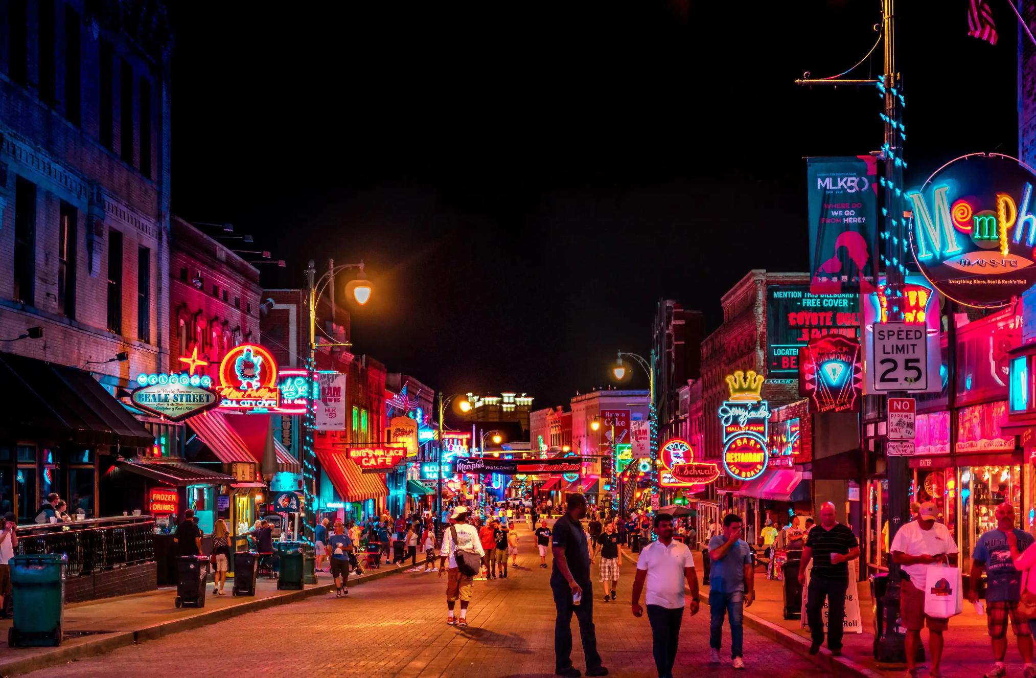 Vibrant night scene on Beale Street in Memphis, with neon signs glowing above blues clubs, bars, and live music venues, and crowds of people walking along the historic street