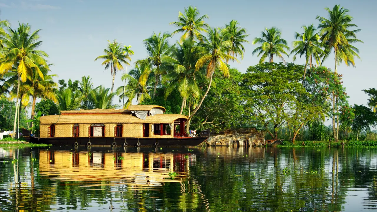 A light brown wicker houseboat on calm water in Kerala, with palm trees on the banks and clear blue skies overhead
