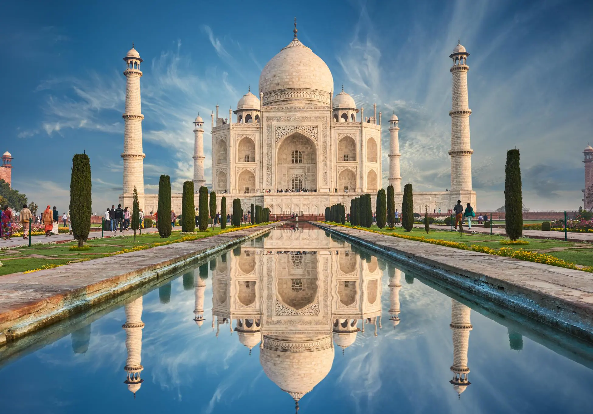 The Taj Mahal reflected in the decorative pool in front of it, surrounded by landscaped gardens and manicured topiaries, under a bright sky with a few clouds