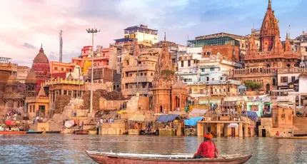 A boatman rowing on the River Ganges in Varanasi, facing the colourful temples and crowded ghats along the ancient city’s waterfront under a soft pastel sky