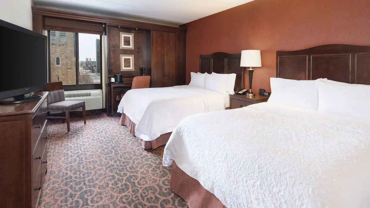 A twin room at Hampton Inn Downtown, Cleveland, featuring two double beds with crisp white linens, dark wood furnishings, warm-toned décor, and a patterned carpet