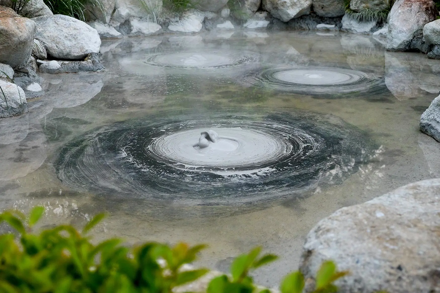 Bubbling grey mud pools in a steamy hot spring at the Mud Hell in Beppu, Japan, surrounded by rocks and greenery