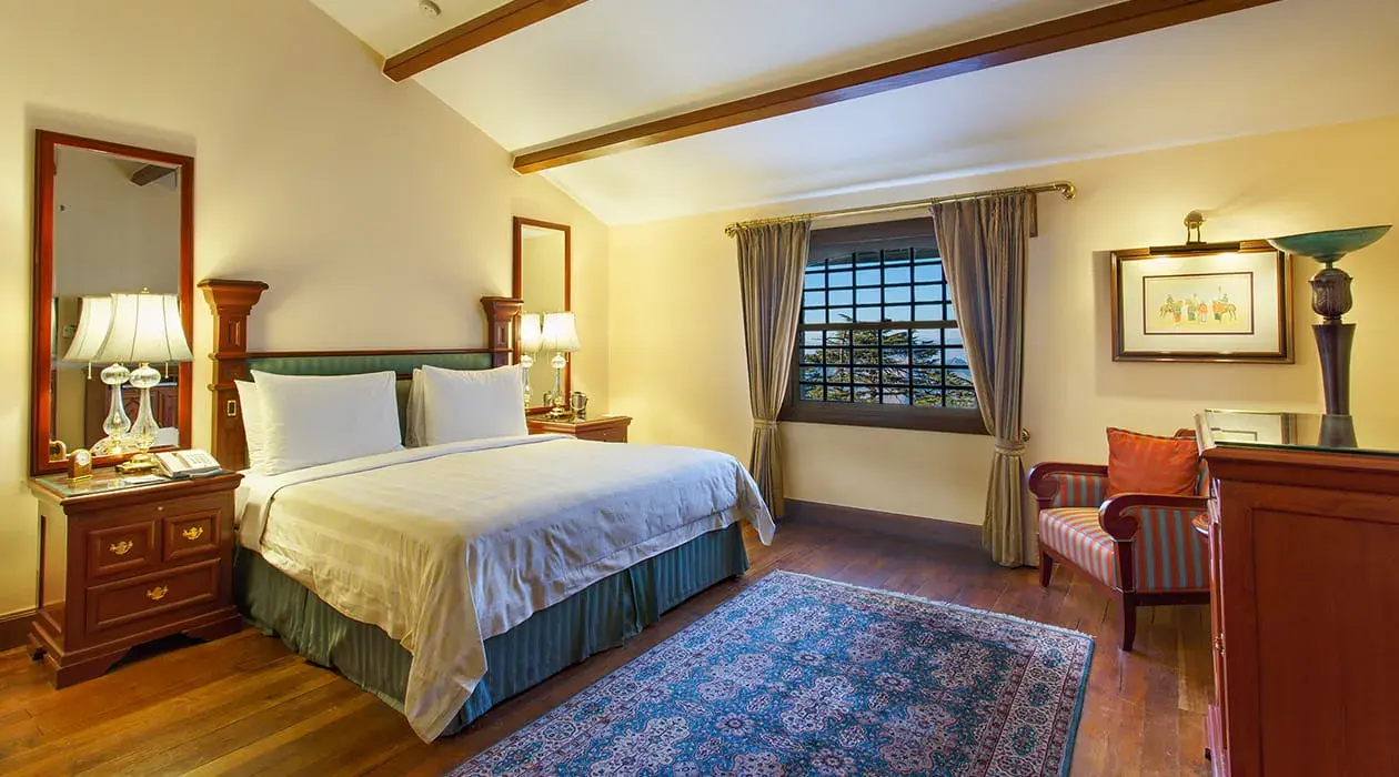 A double room at the Oberoi Cecil hotel in Shimla, featuring wooden floors, a blue patterned rug, cosy warm furnishings, and a window with views of the surrounding trees