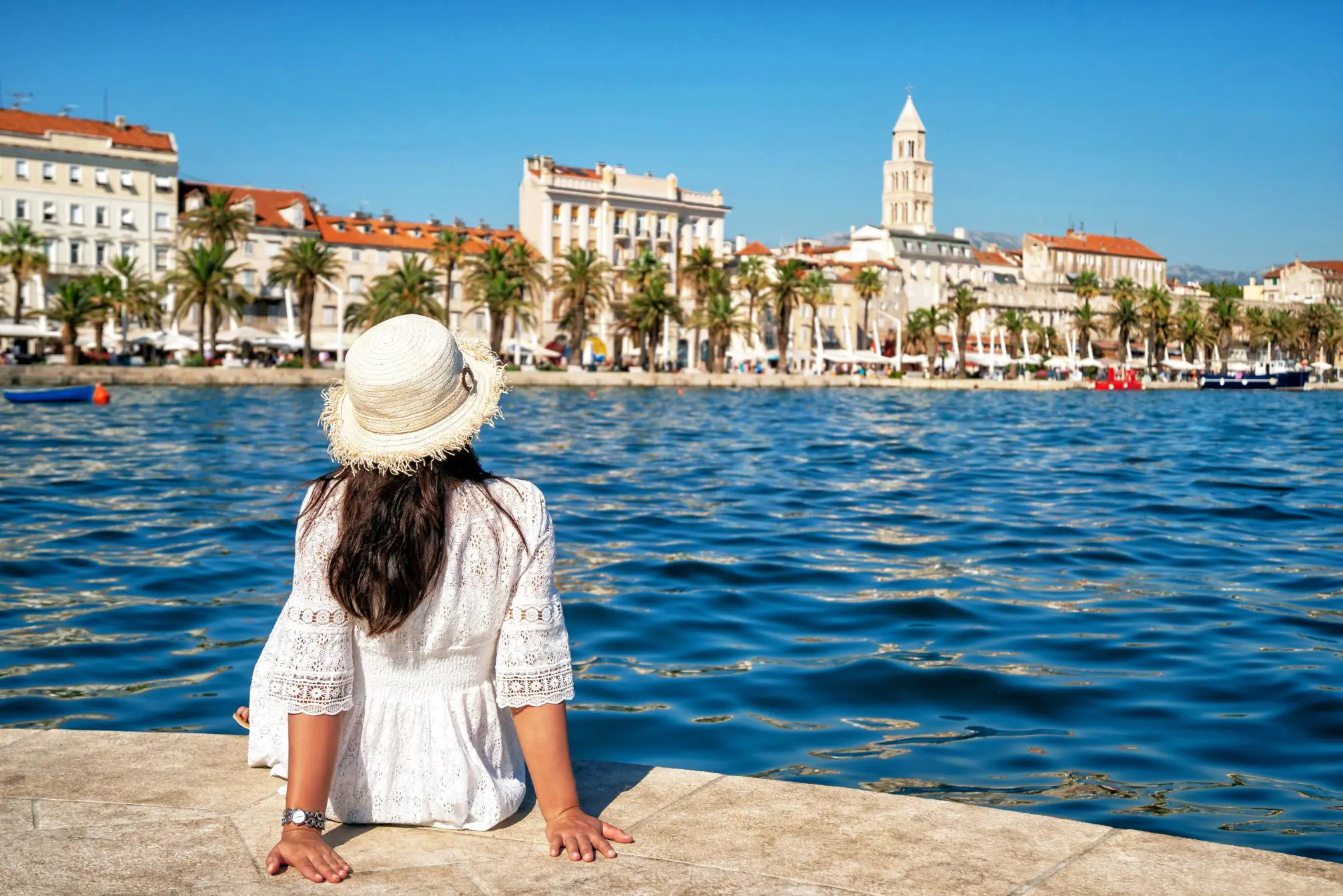 A woman in a white dress and straw hat sitting on the harbour edge in Split, Croatia, looking across the calm Adriatic Sea toward the historic waterfront and the bell tower of St. Domnius Cathedral