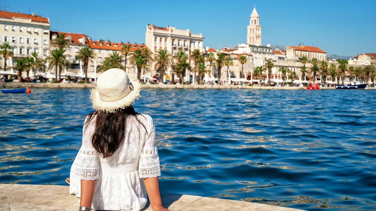 A woman in a white dress and straw hat sitting on the harbour edge in Split, Croatia, looking across the calm Adriatic Sea toward the historic waterfront and the bell tower of St. Domnius Cathedral
