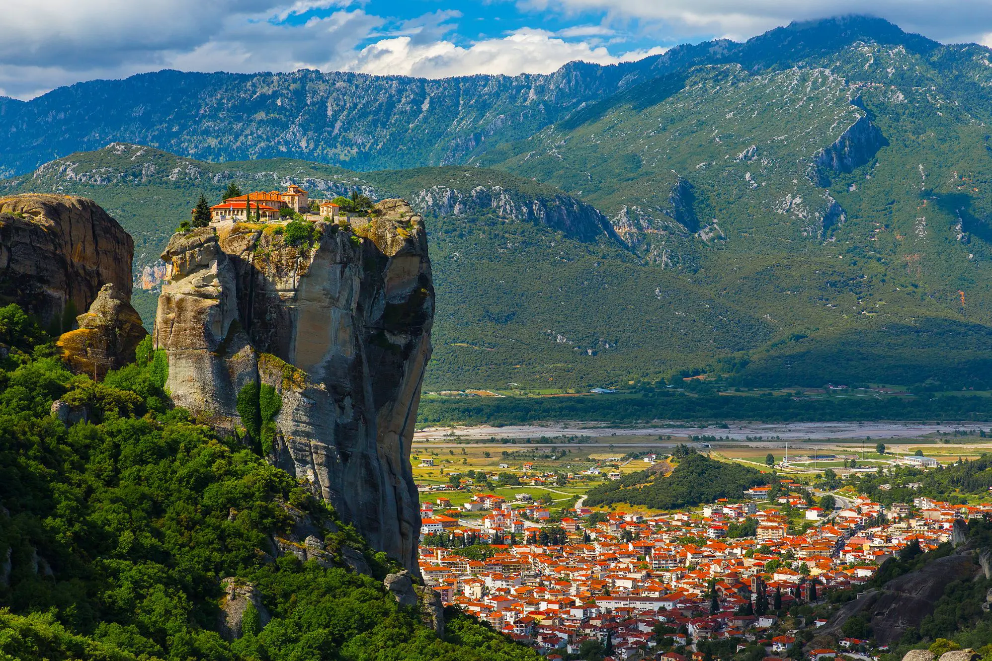 High rock precipice with a monastry on the top. In the background, mountains and a town on the land below 