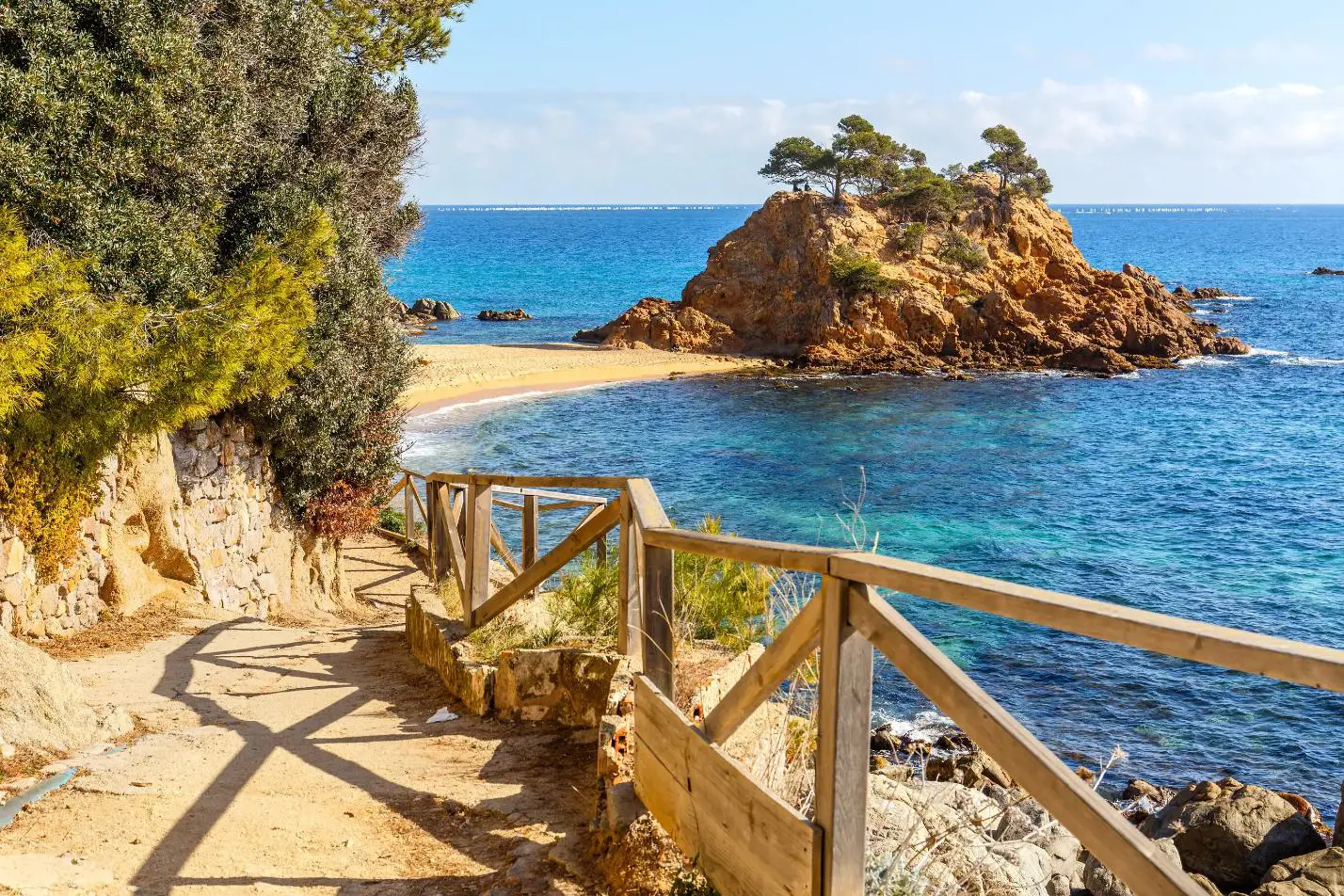 Scenic view of the Cami de Ronda coastal path along Costa Brava, winding above rugged cliffs and turquoise Mediterranean waters, with pine trees and rocky coves dotting the shoreline