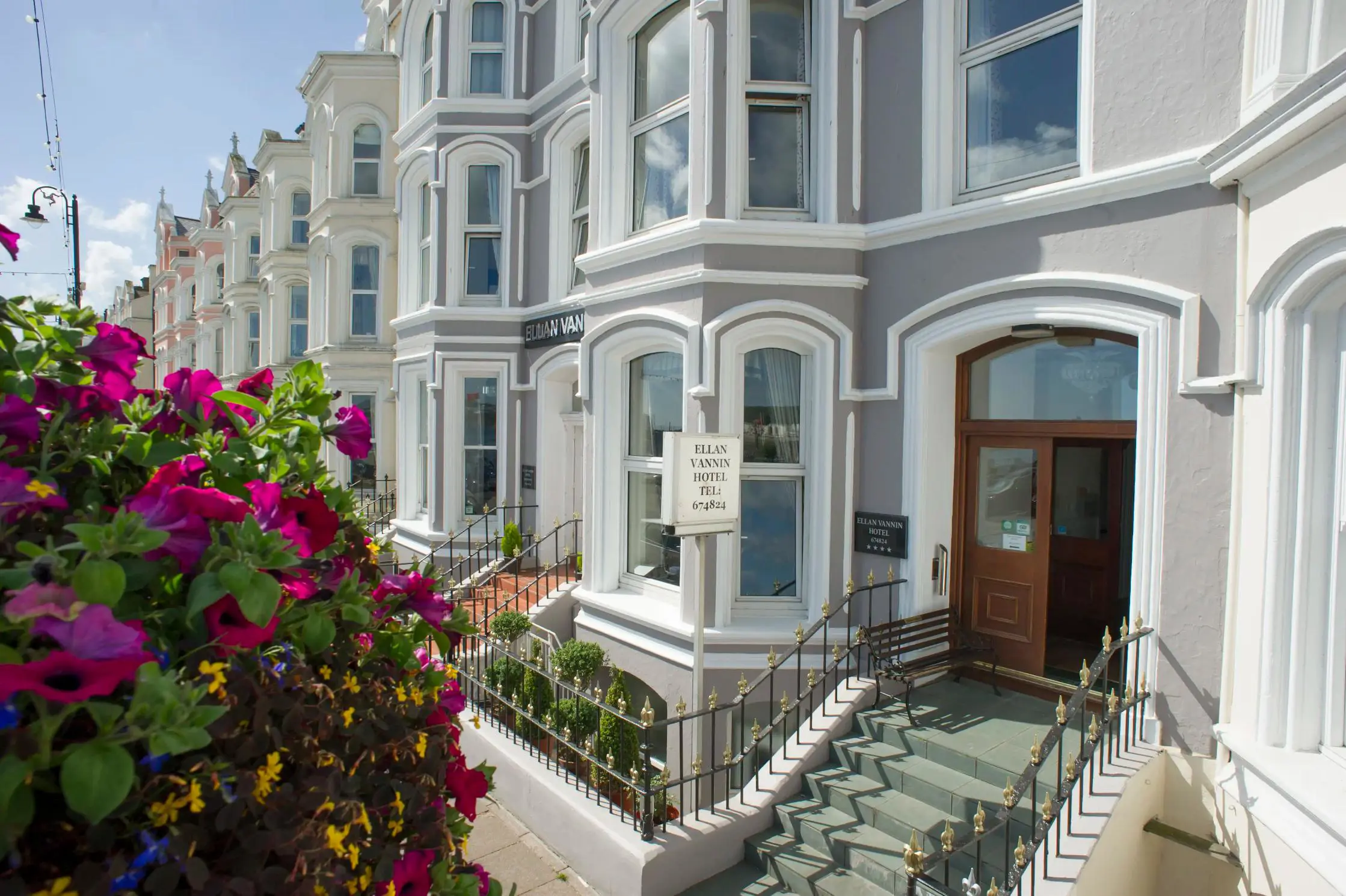 Front entrance of the Ellan Vannin Hotel, a traditional building with bay windows, grey and white exterior, decorative railings, and vibrant purple flowers in the foreground