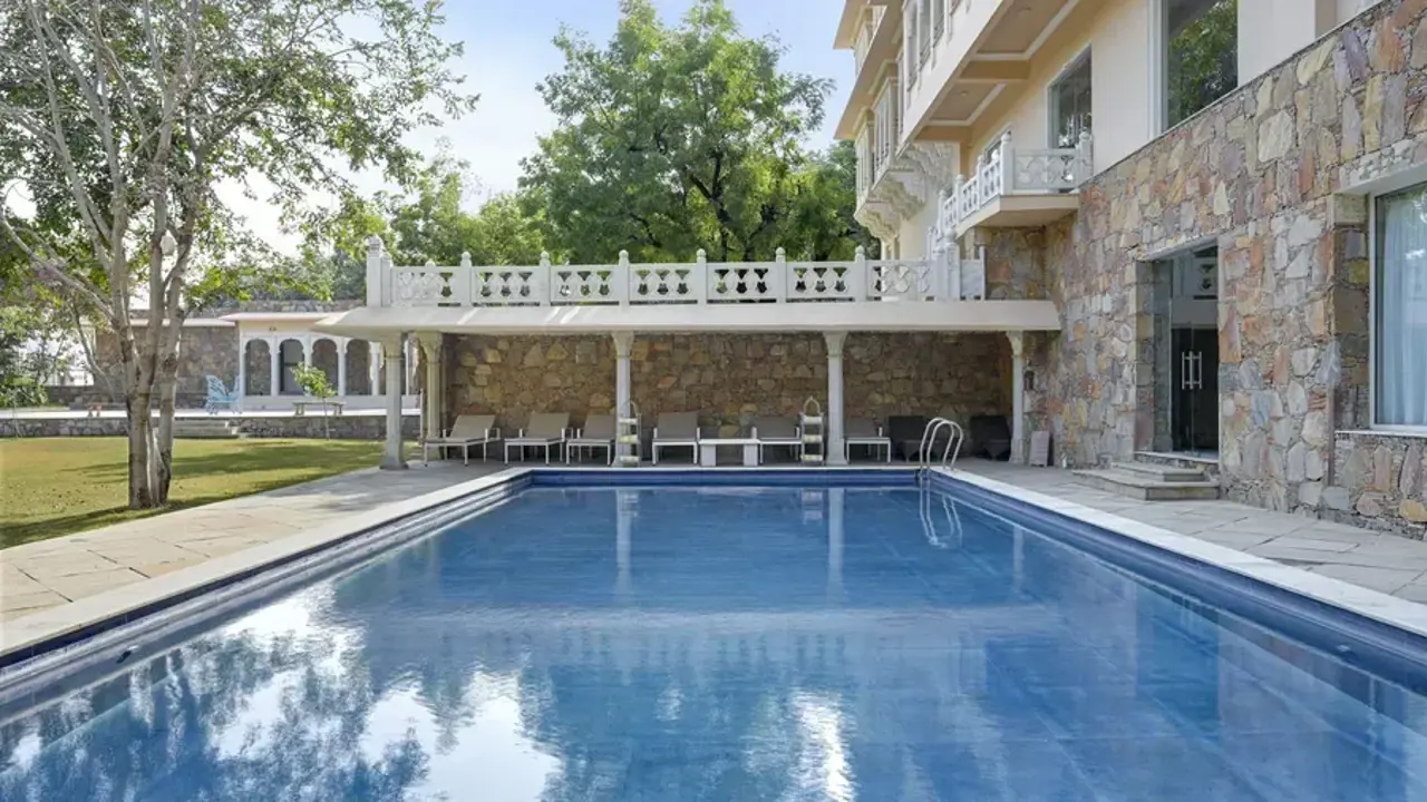 Outdoor swimming pool at the Fateh Niwas Hotel, beside a stone building, with shaded seating and trees in the background