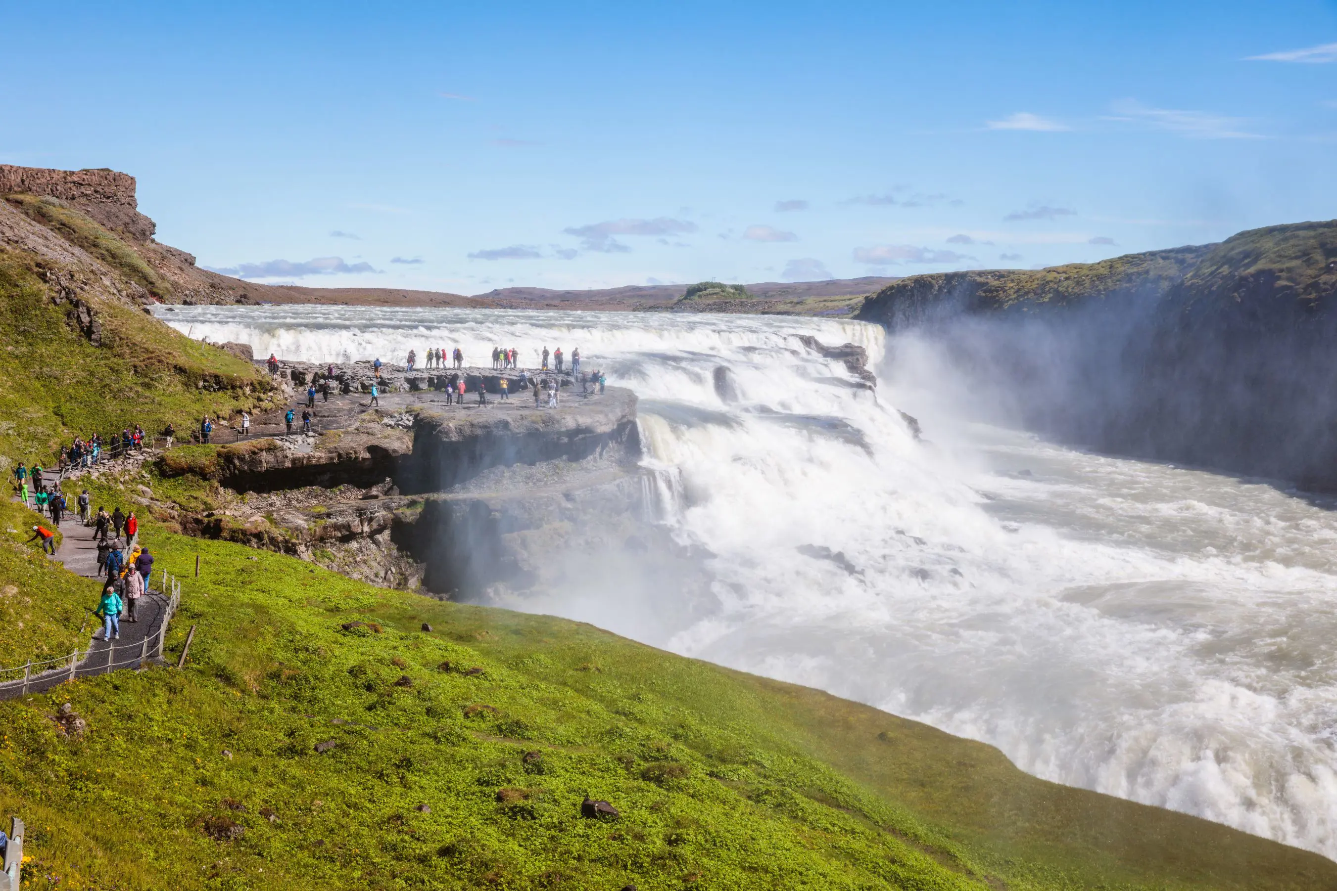 High angle shot of the mouth of a waterfall. People stood on the rock next to it, grassy land in the forefront