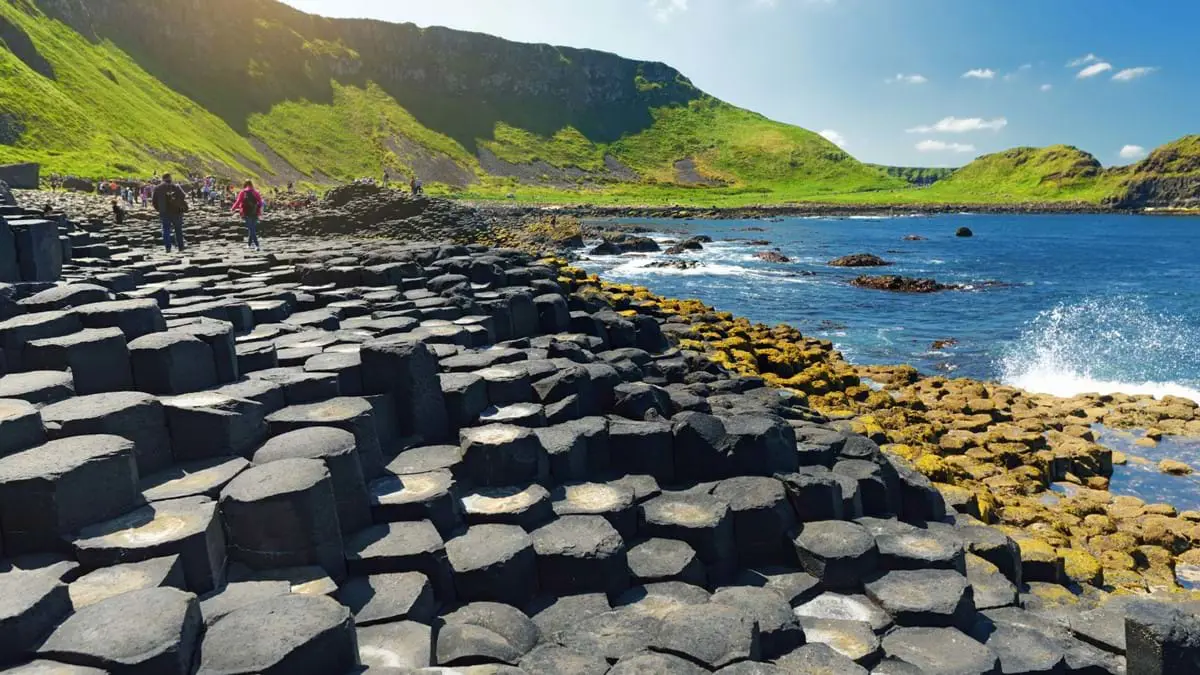 Large stones of the Giant's Causeway at the forefront, with waves crashing into them, back of people in the distance walking towards a grassy mountain