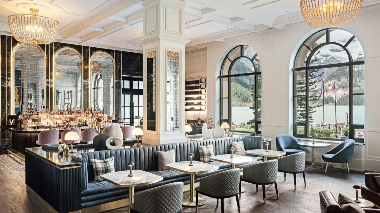 Elegant, brightly lit, bar area at Fairmont Chateau, Lake Louise featuring a stylish seating area, a modern bar in the background, and large arched windows with a view of Lake Louise