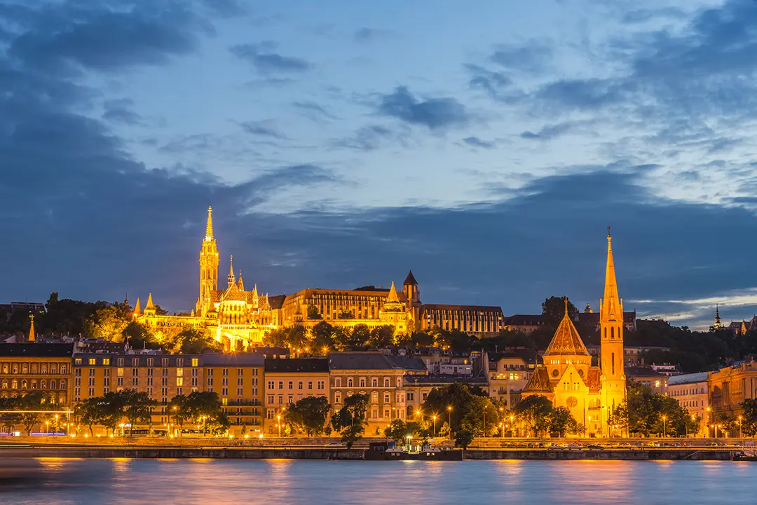 View of skyline of Buda, from Pest. With lit up buildings including two churches with spiked turrets.