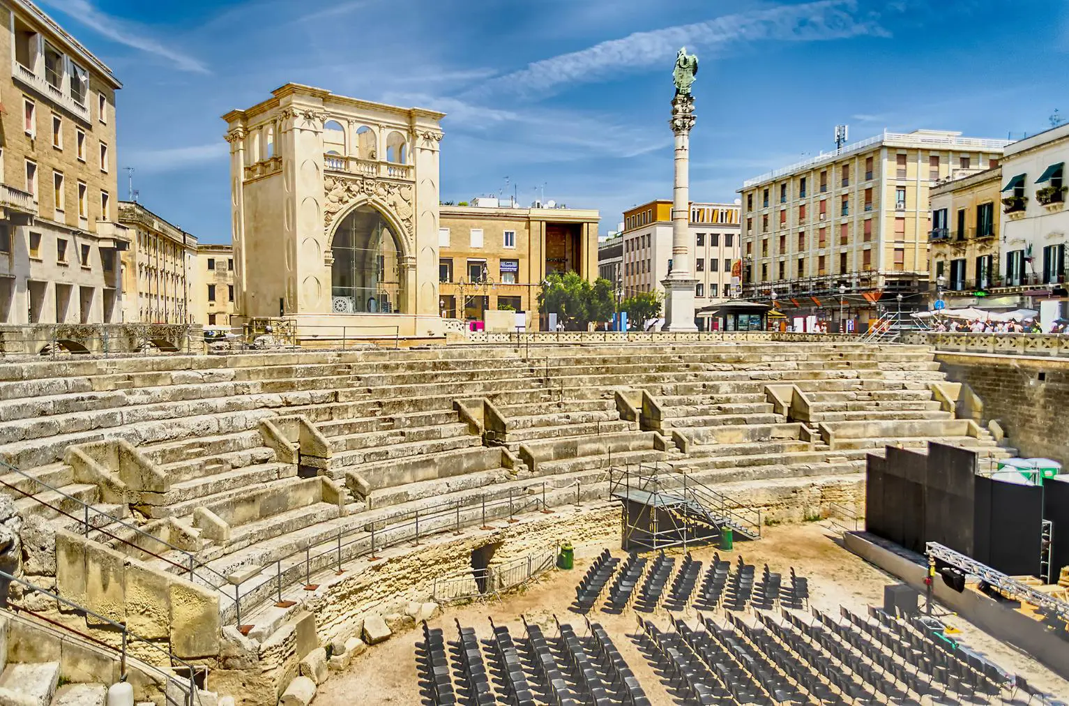 A Roman amphitheatre with stone seating in Sant'Oronzo Square, Lecce, under a clear blue sky, surrounded by modern buildings and event chairs on the ground