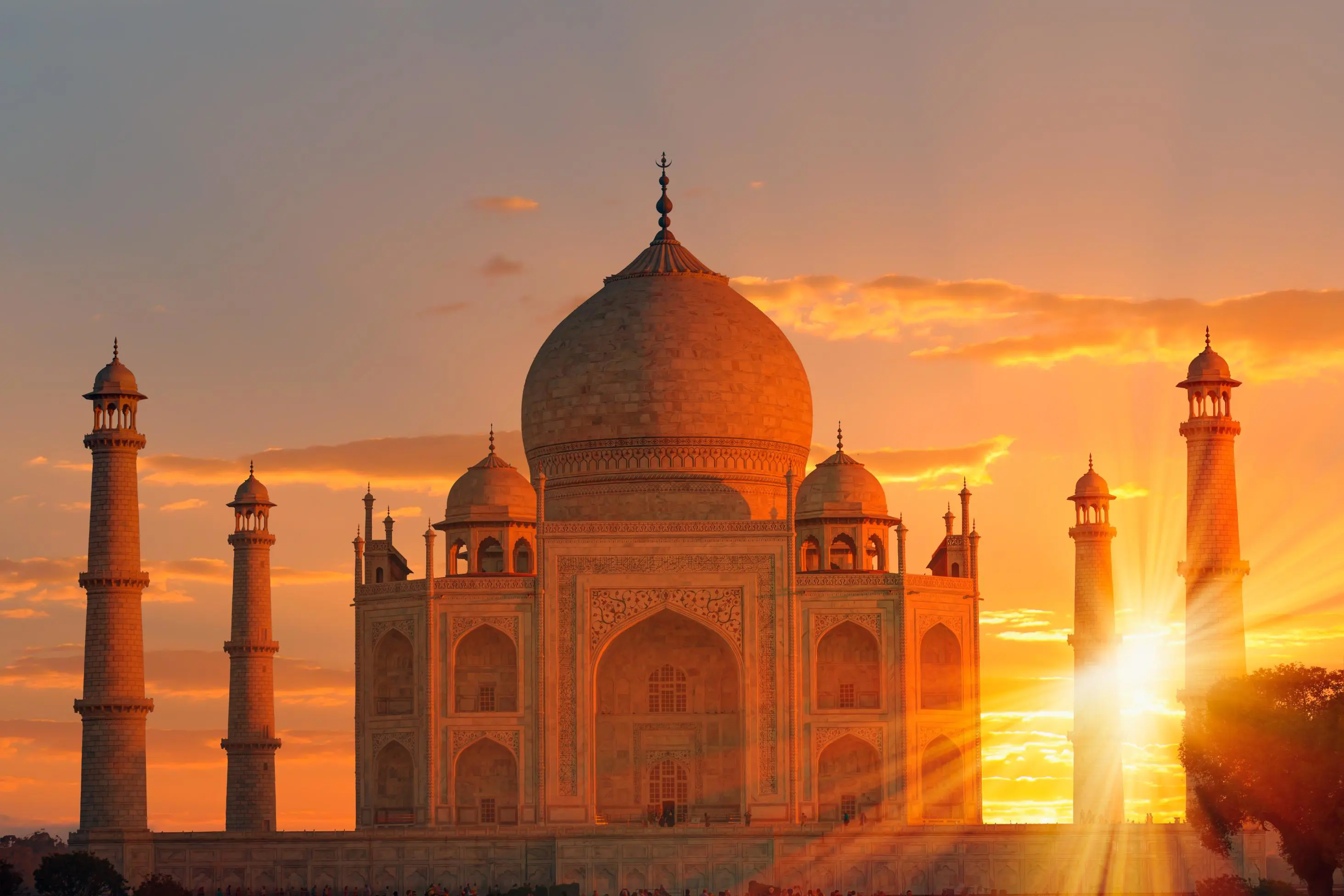 The Taj Mahal bathed in golden sunlight at sunset, with the sun setting between its elegant minarets, highlighting the detailed marble carvings and domed architecture