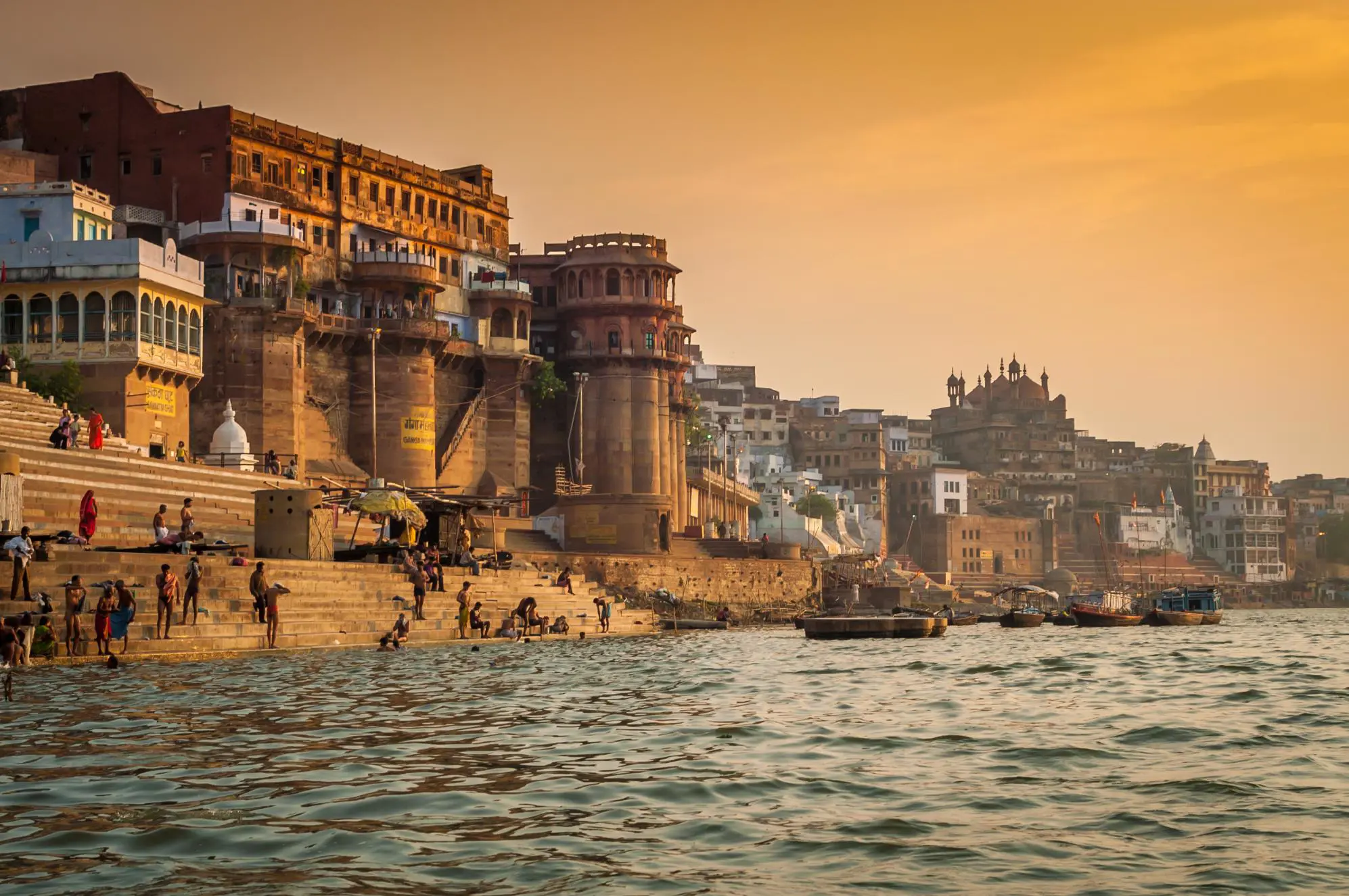 People gathering and bathing along the ghats of the River Ganges in Varanasi during sunset, with historic stone buildings and temples rising along the riverfront