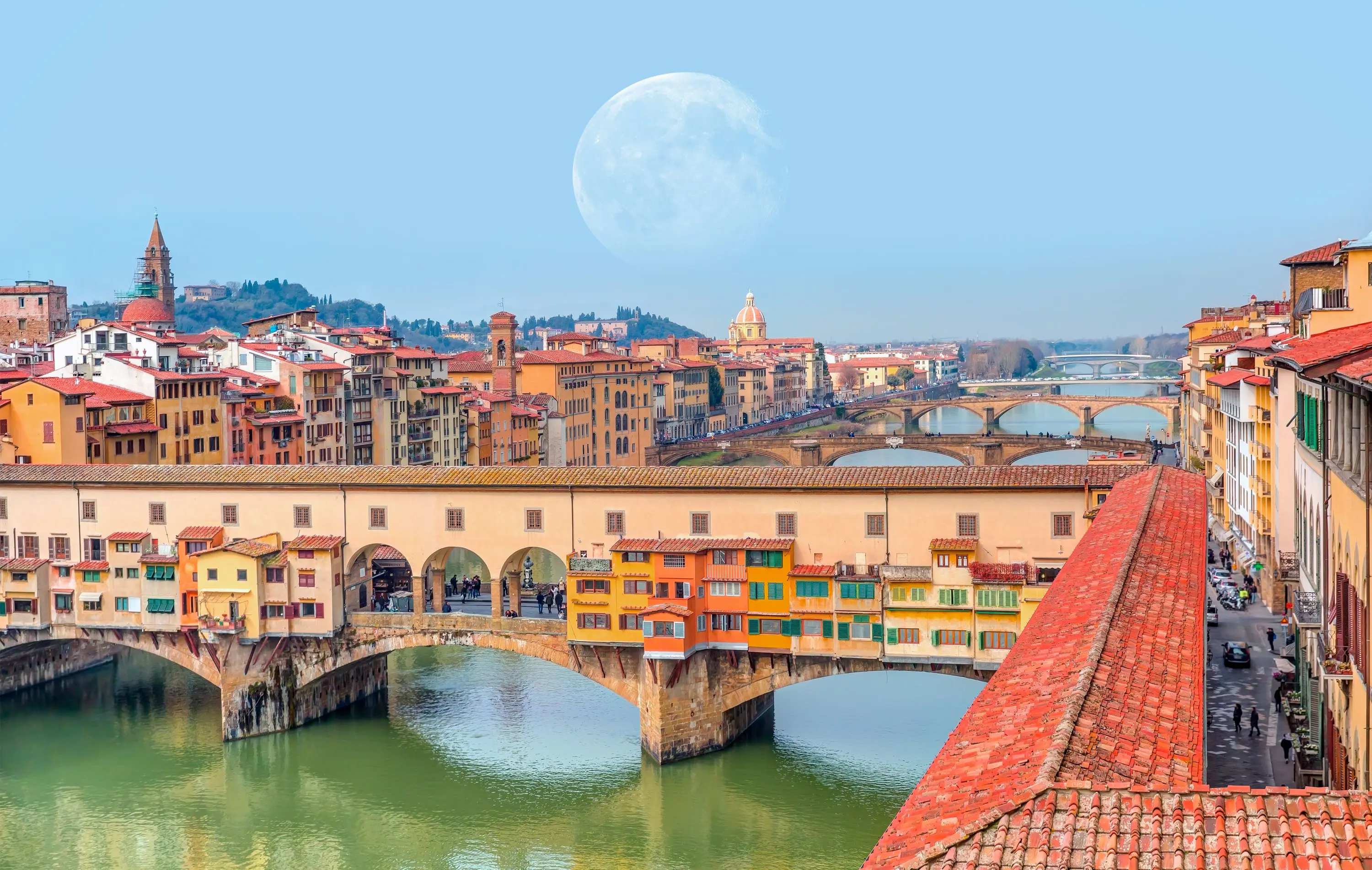 View of Ponte Vecchio Bridge showing buildings, the rest of The Arno River in the background, and a faint full moon in the sky