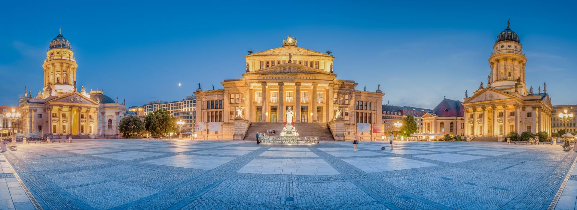 Wide angle shot of a grand, lit up building, with a building either side, both with a tower with a domed top. The middle building has a white statue of a person on a raised platform in front. The sky is a bright blue.