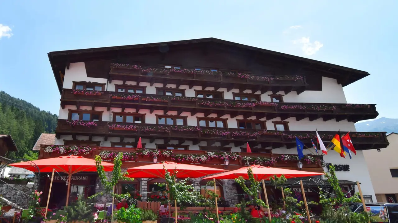 A traditional alpine-style hotel in Austria with wooden balconies decorated with pink and white flowers. Red parasols shade an outdoor café area which is surrounded by greenery, and several international flags are displayed at the front