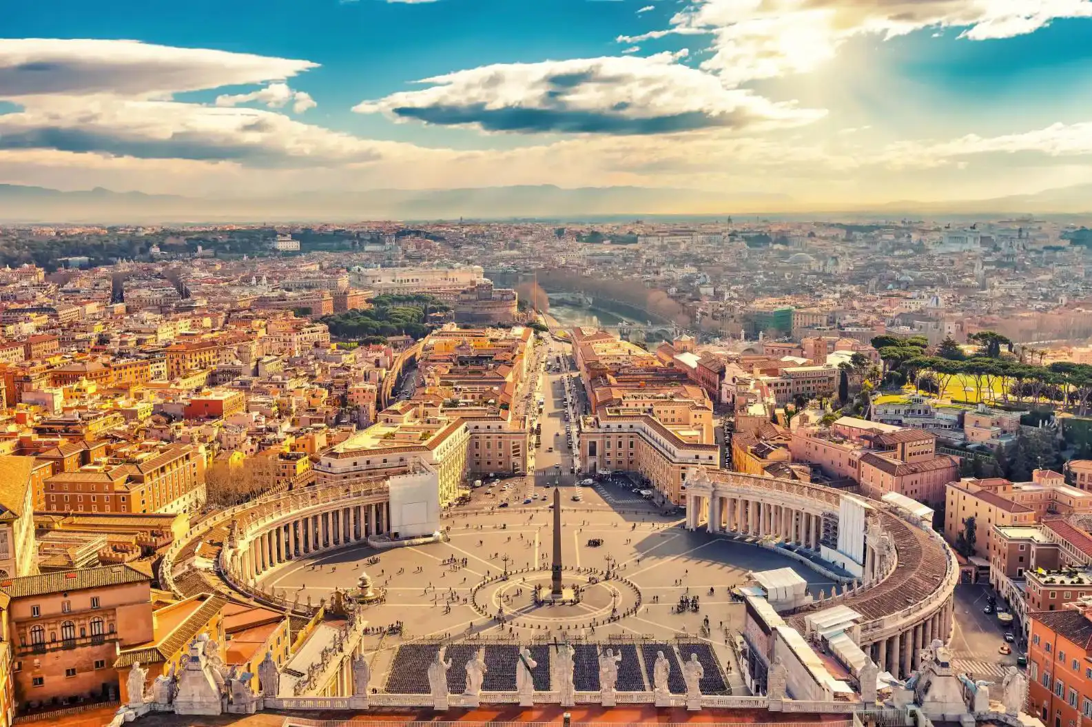Aerial view of St. Peter’s Square in Vatican City, bathed in golden sunlight with Rome in the distance under a partly cloudy sky