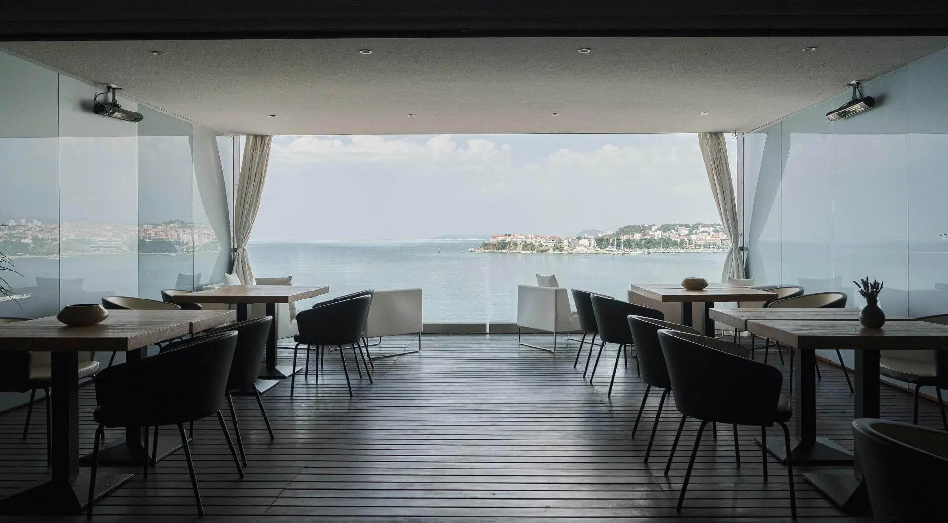 A stylish indoor restaurant with minimalist wooden tables and black chairs, looking out through glass windows onto a stunning coastal view of Podstrana and the Adriatic Sea