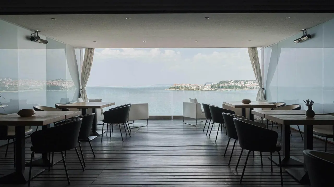 A stylish indoor restaurant with minimalist wooden tables and black chairs, looking out through glass windows onto a stunning coastal view of Podstrana and the Adriatic Sea