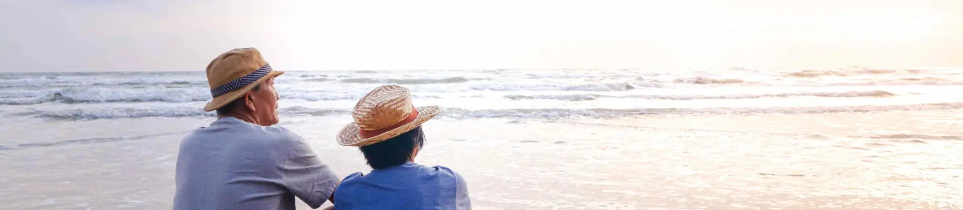 An elderly couple in straw hats sits side by side on a sandy beach, peacefully watching the calm ocean as the sun sets