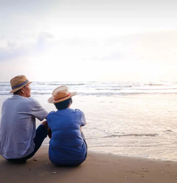 An elderly couple in straw hats sits side by side on a sandy beach, peacefully watching the calm ocean as the sun sets