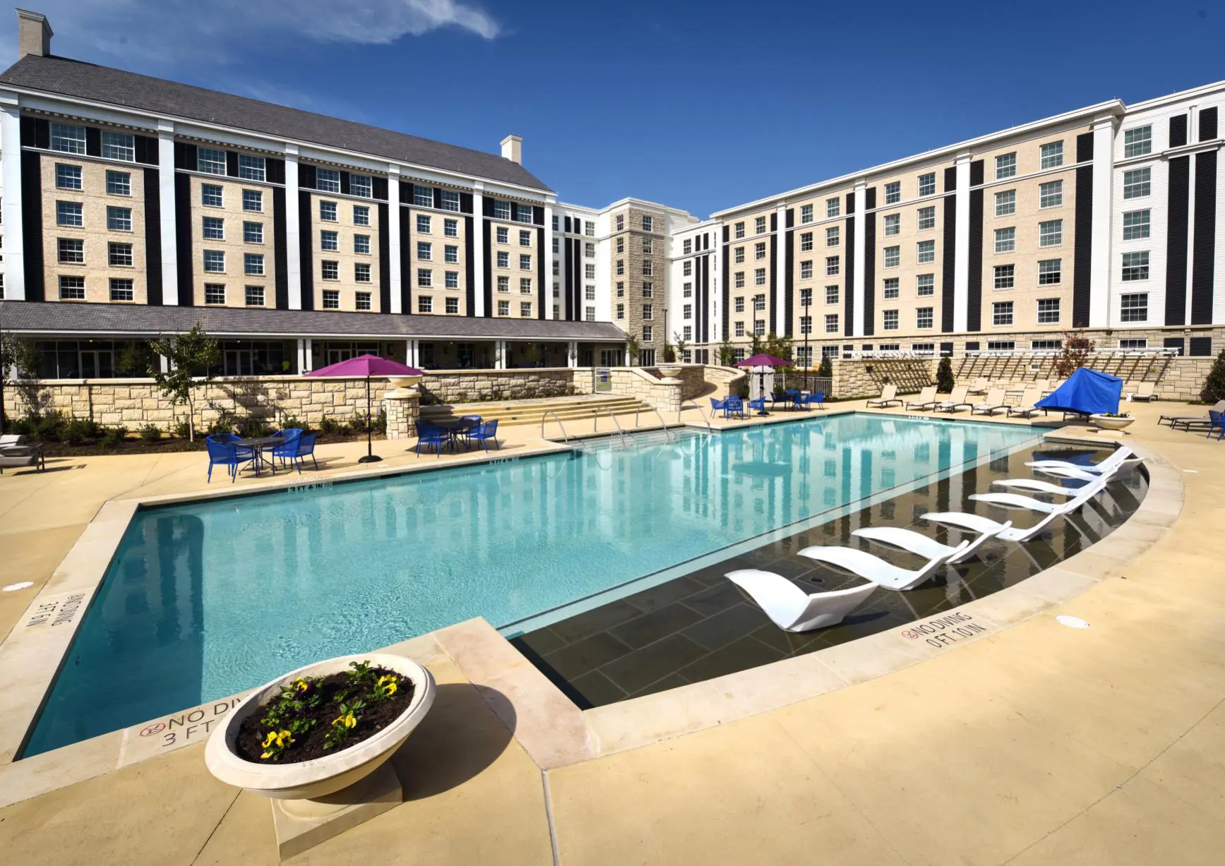 Outdoor pool at The Guest House at Graceland in Memphis, Tennessee, with in-water loungers, parasols, and the hotel’s brick building in the background under a clear sky