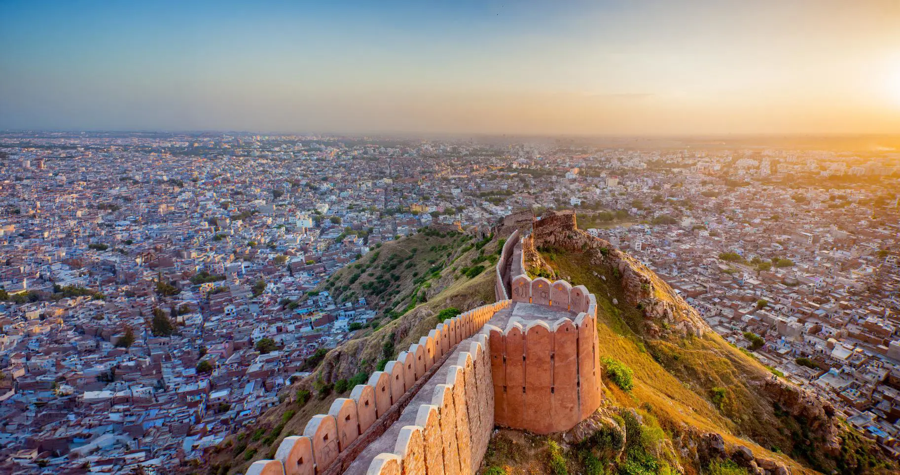 An aerial view of Nahargarh Fort in Jaipur stretching along a ridge at sunset, overlooking the sprawling cityscape below under a hazy orange sky