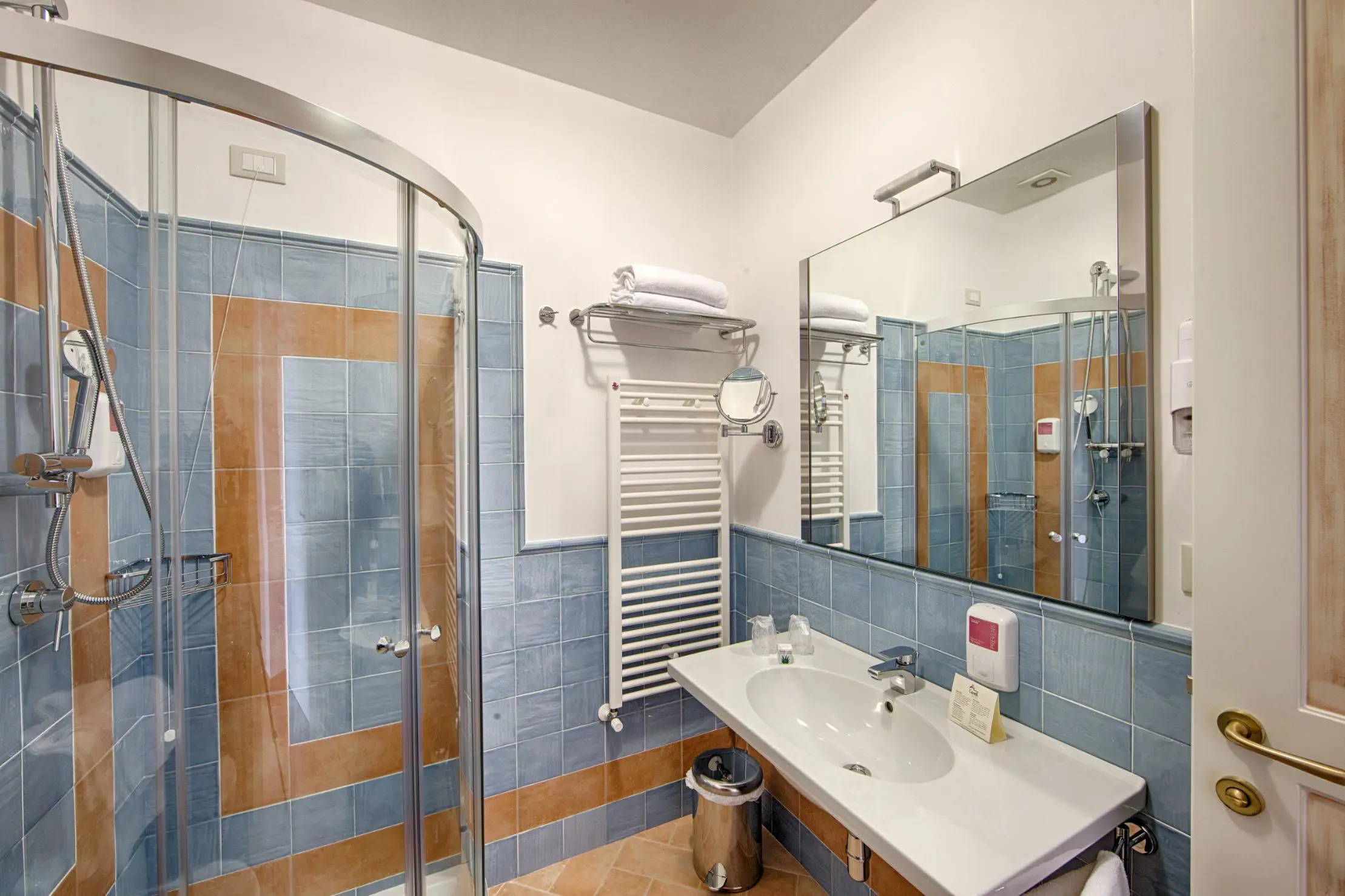 Modern bathroom at Masseria Casselli Hotel with blue and beige tiled walls, a curved glass shower enclosure, a white sink with a large mirror and a towel rack