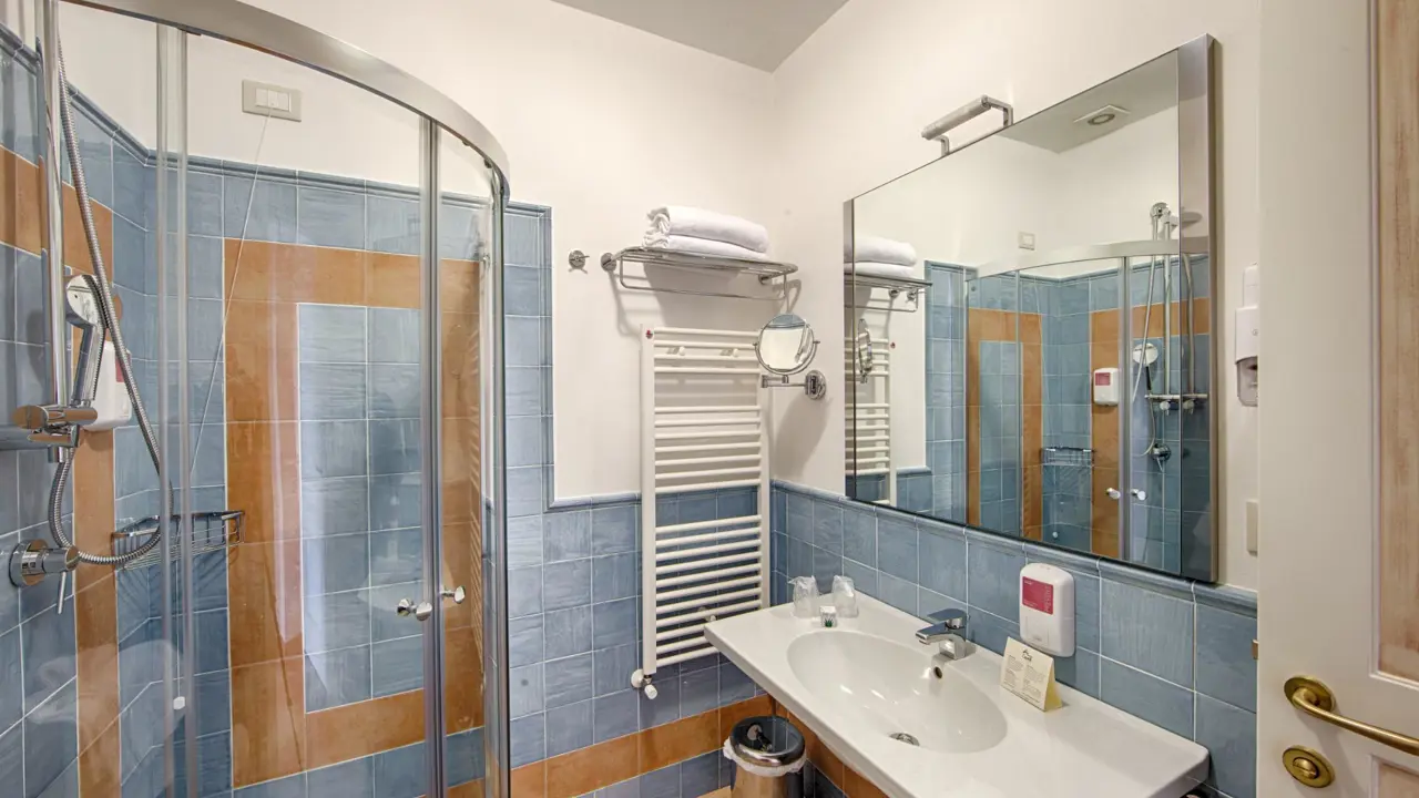 Modern bathroom at Masseria Casselli Hotel with blue and beige tiled walls, a curved glass shower enclosure, a white sink with a large mirror and a towel rack