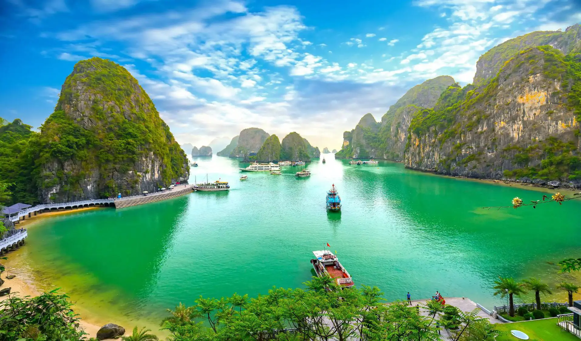 Panoramic view of Ha Long Bay in Vietnam, with emerald green waters dotted with traditional wooden boats, surrounded by towering limestone karsts and lush, forested islets under a partly cloudy sky