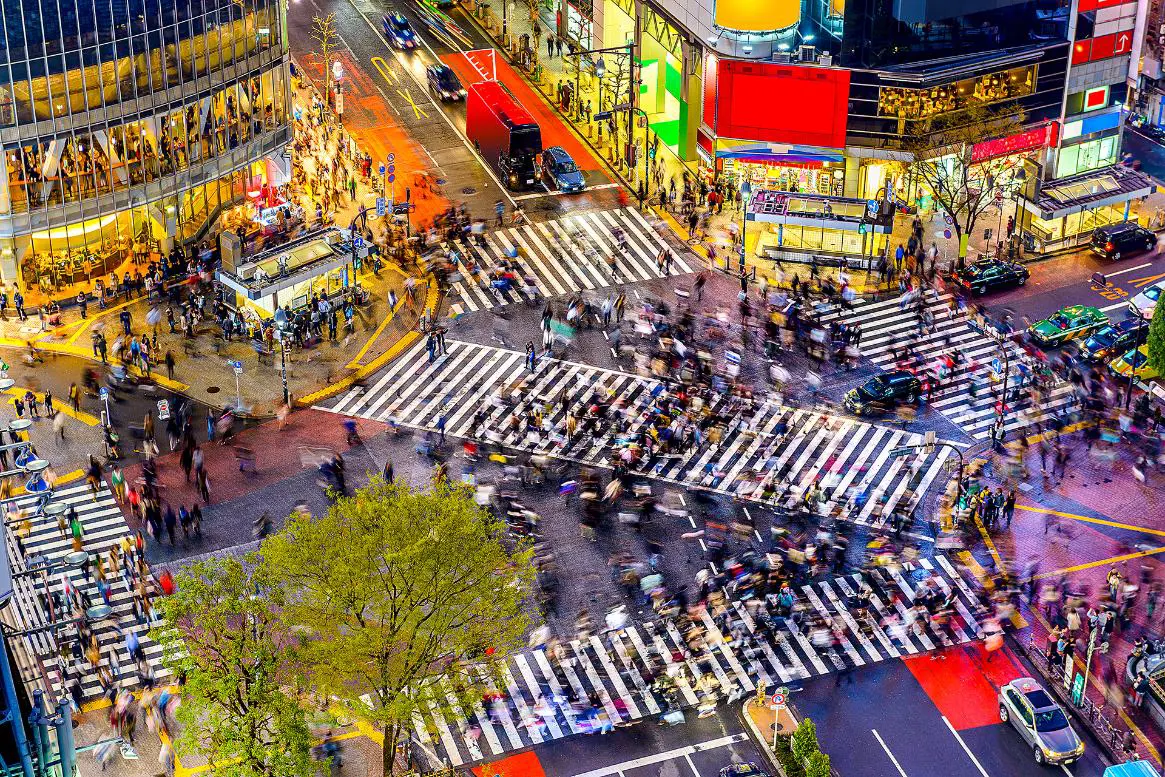 A vibrant view of Shibuya Crossing in Tokyo at night, bustling with crowds of pedestrians crossing from all directions, surrounded by bright billboards and colourful neon signs illuminating the streets