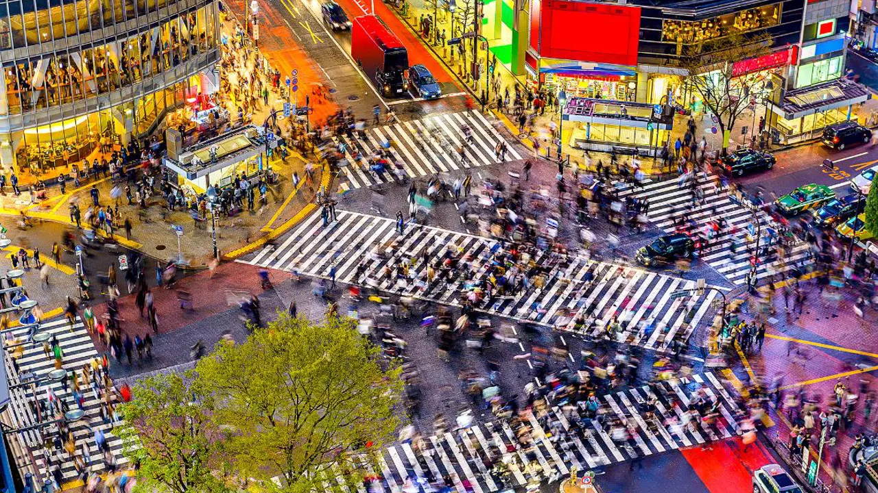 A vibrant view of Shibuya Crossing in Tokyo at night, bustling with crowds of pedestrians crossing from all directions, surrounded by bright billboards and colourful neon signs illuminating the streets