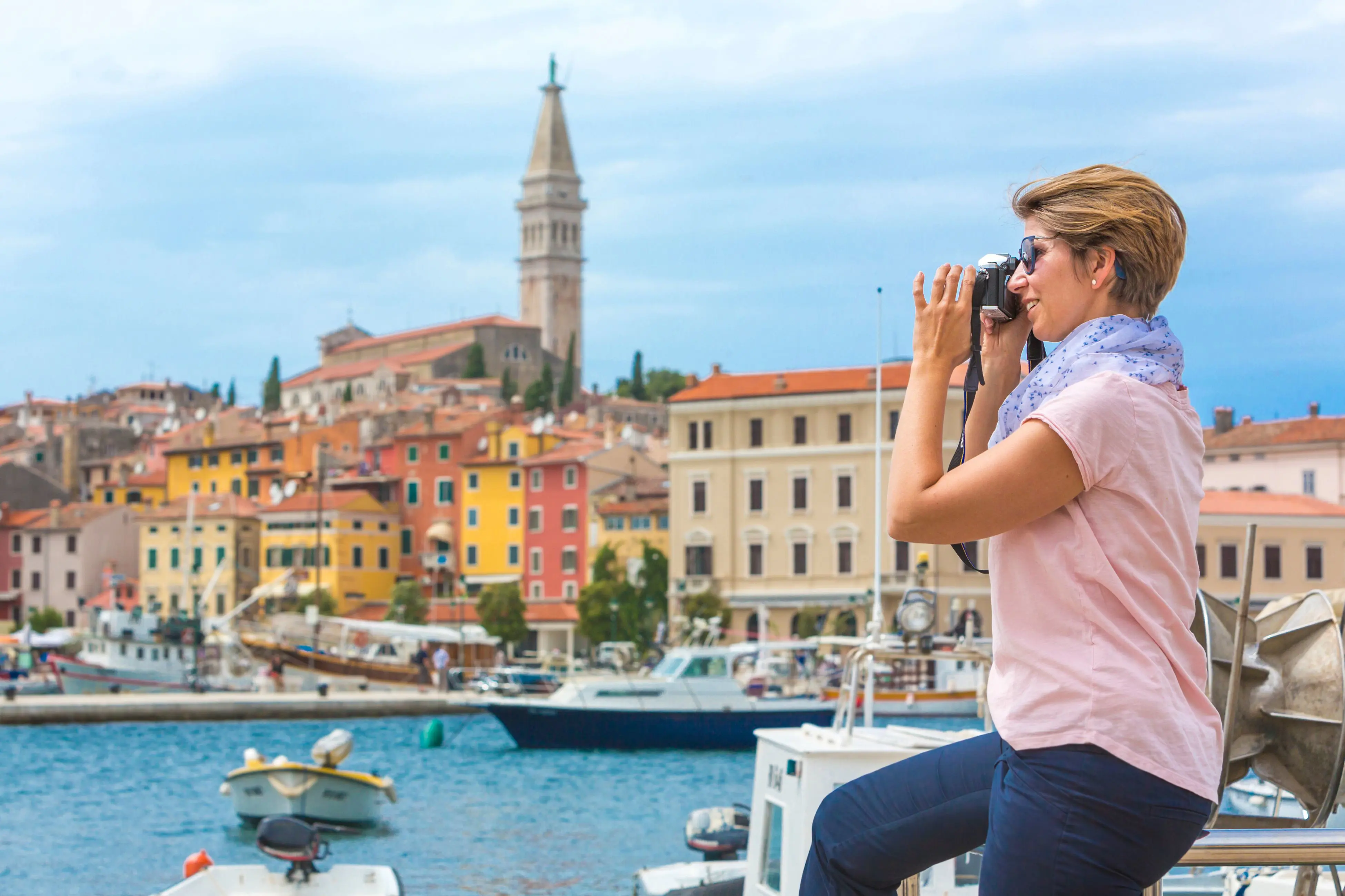 Shot a woman facing the left, taking a picture on a large camera with water and boats docked next to her. Red, orange and yellow buildings in the background and a tower behind.
