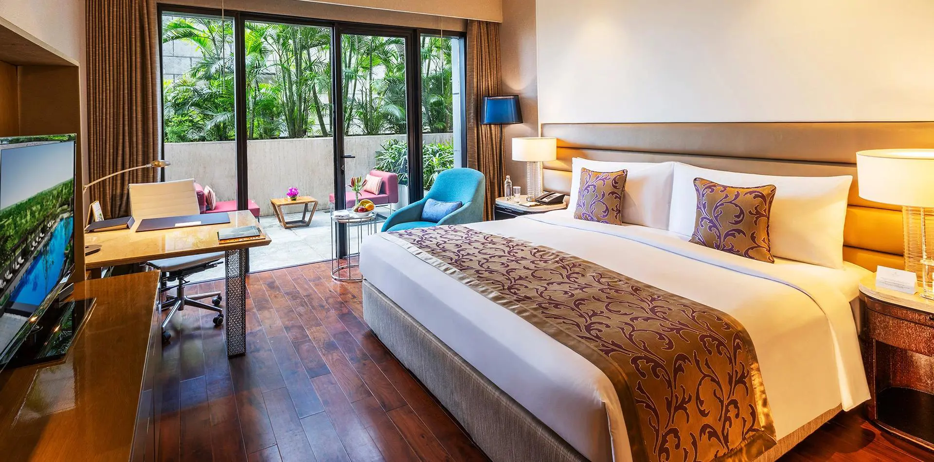Cosy double room at the Leela Ambience Convention Hotel in Delhi, featuring warm lighting, wooden floors, and a terrace outside the patio doors with two pink lounge chairs and a small coffee table