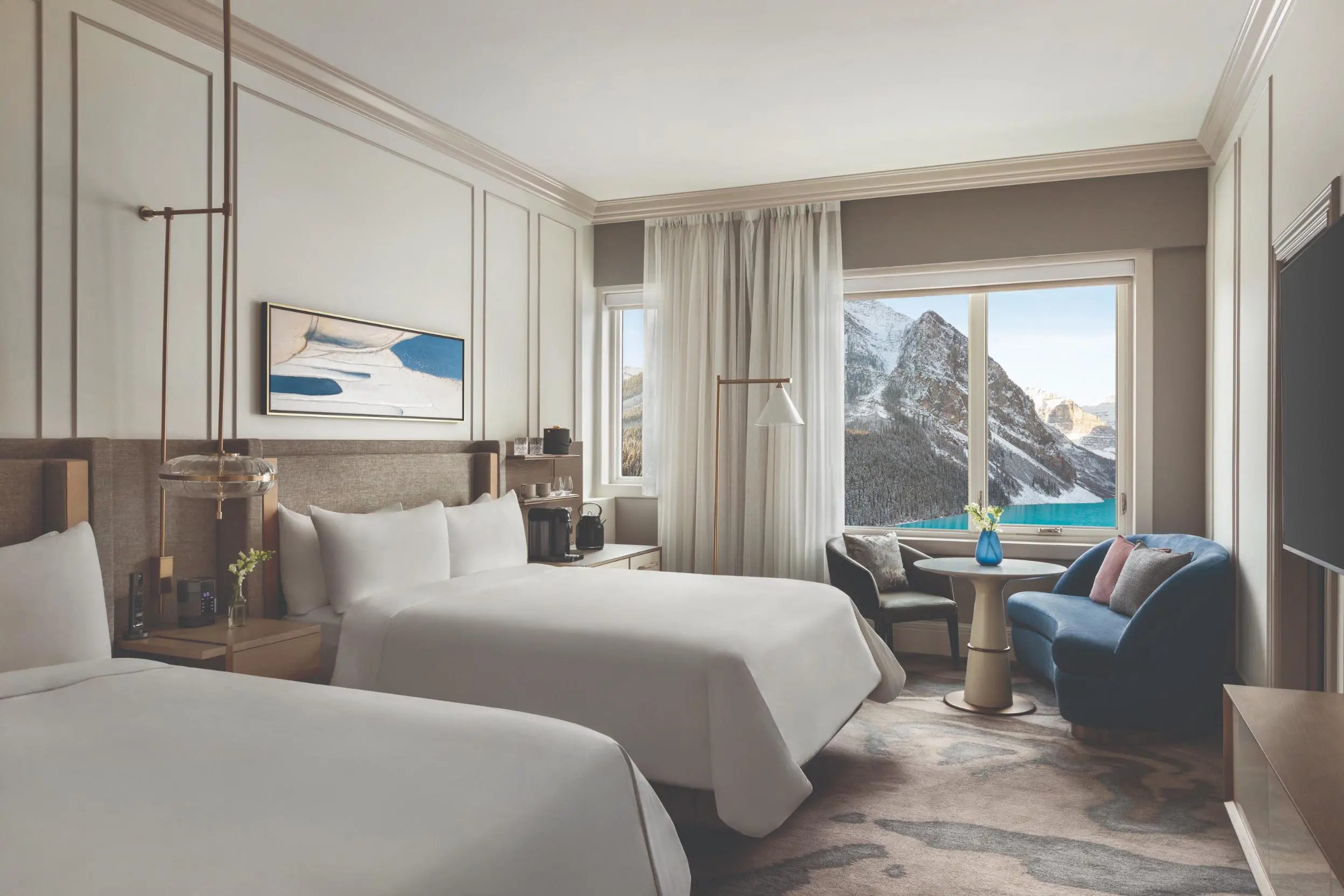 Elegant Fairmont Chateau hotel room with two queen beds, abstract artwork above the headboards, a sitting area with a round table and two chairs, and a window offering views of snow-dusted forested mountains
