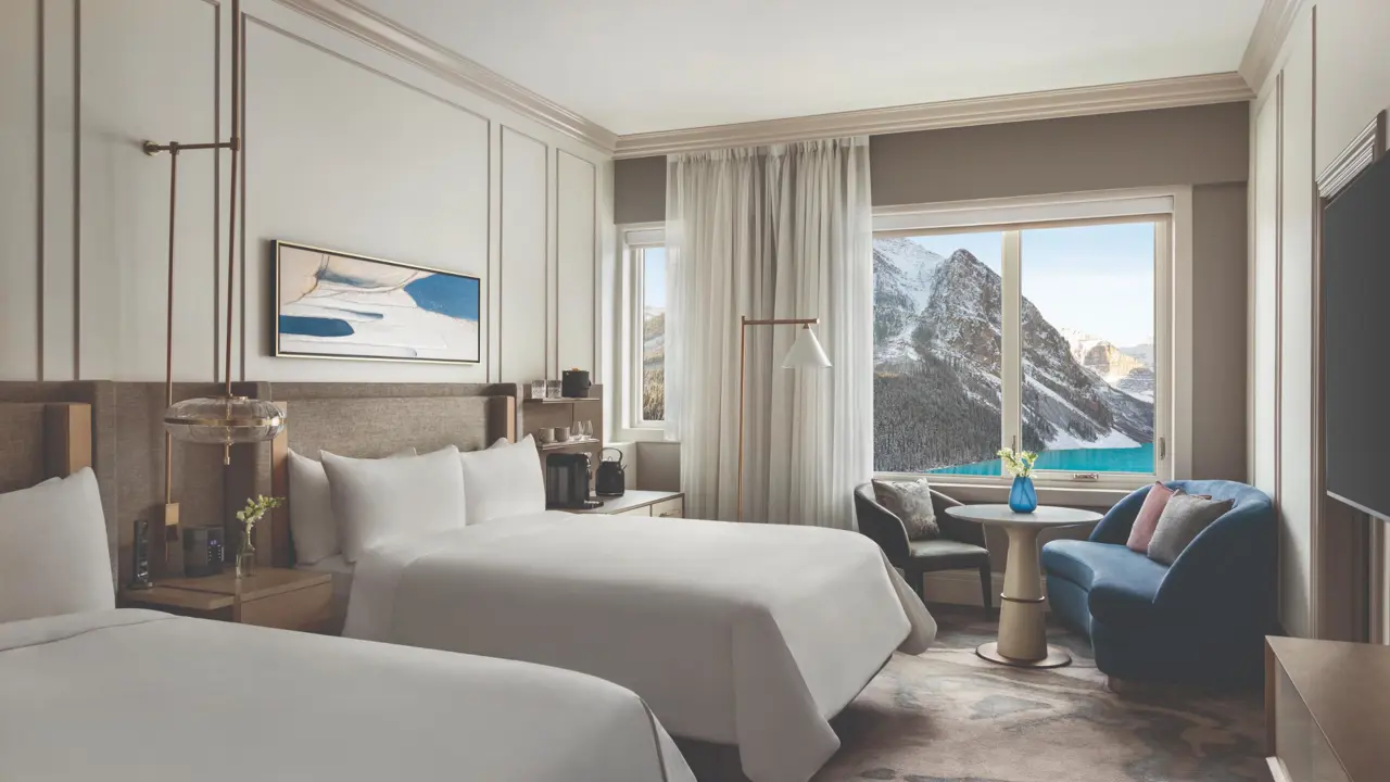 Elegant Fairmont Chateau hotel room with two queen beds, abstract artwork above the headboards, a sitting area with a round table and two chairs, and a window offering views of snow-dusted forested mountains