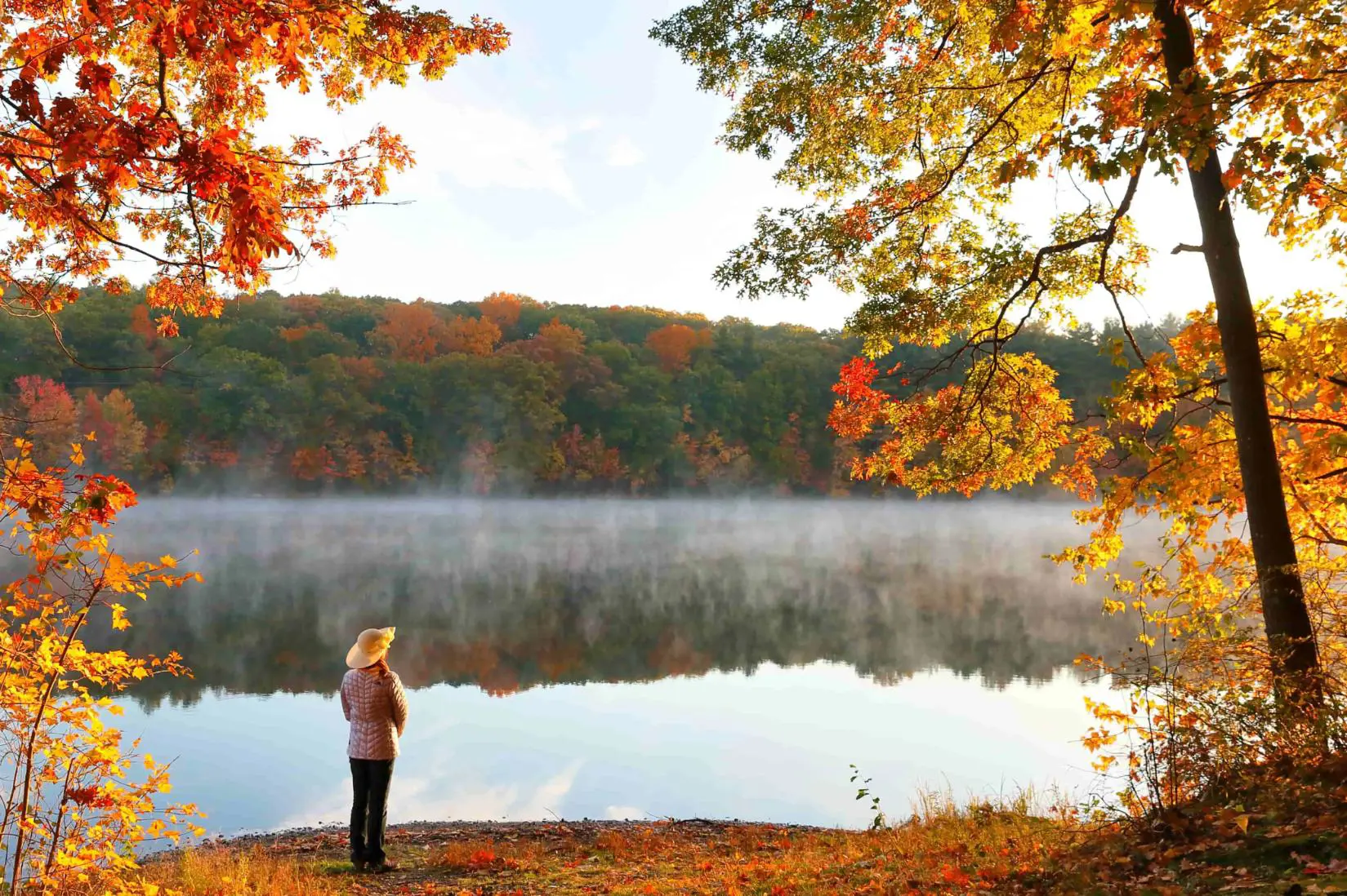 A woman standing by a lake, surrounded by scenic New England autumn foliage with vibrant red, orange, and yellow leaves covering rolling hills